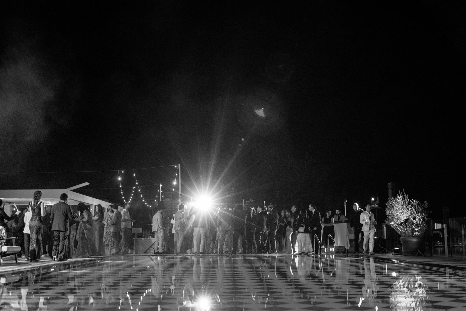 Black and white photo of wedding guests dancing on reflective floor under bright lights.