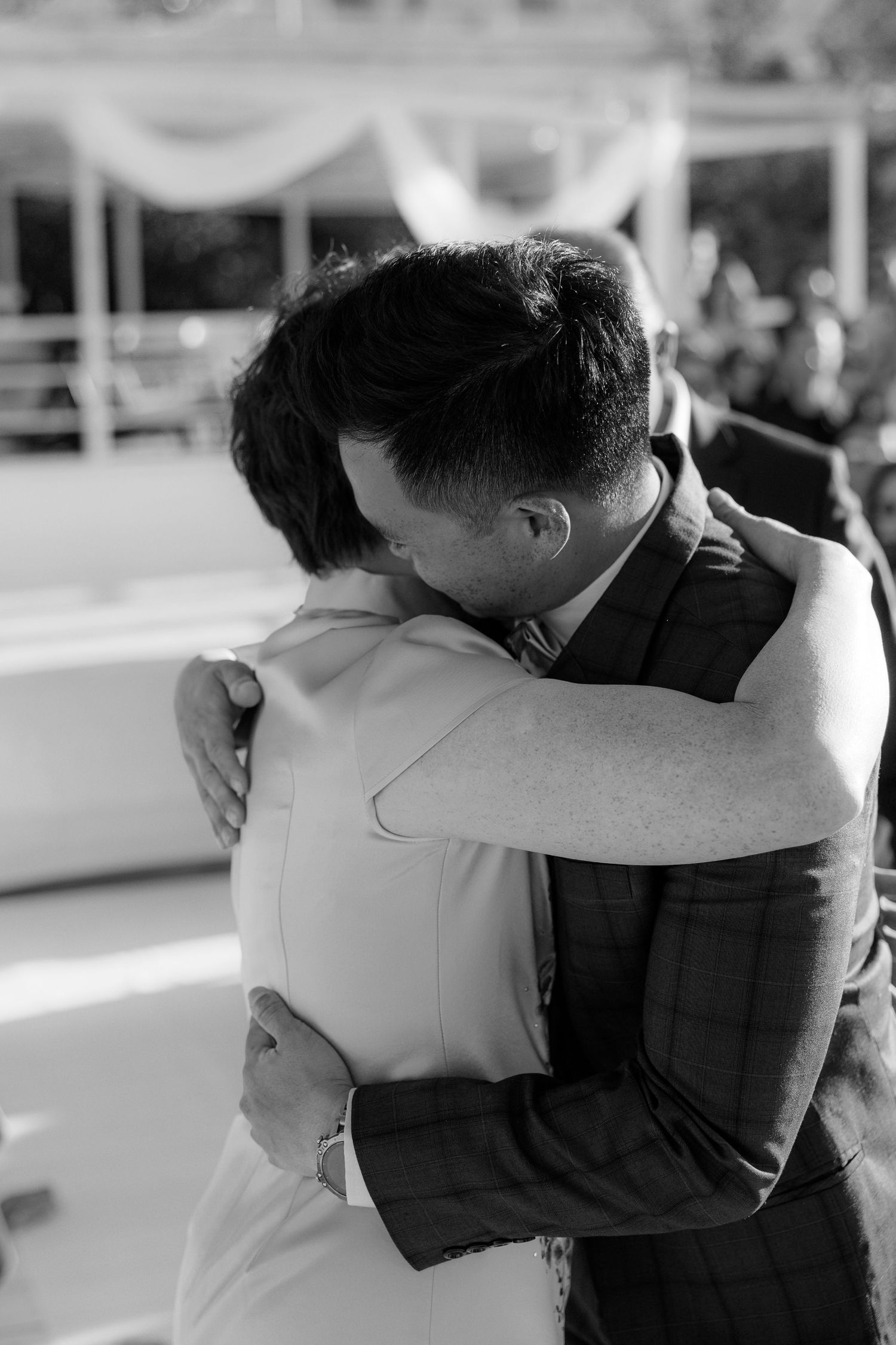 Two men share an emotional embrace in black and white photo at wedding ceremony.