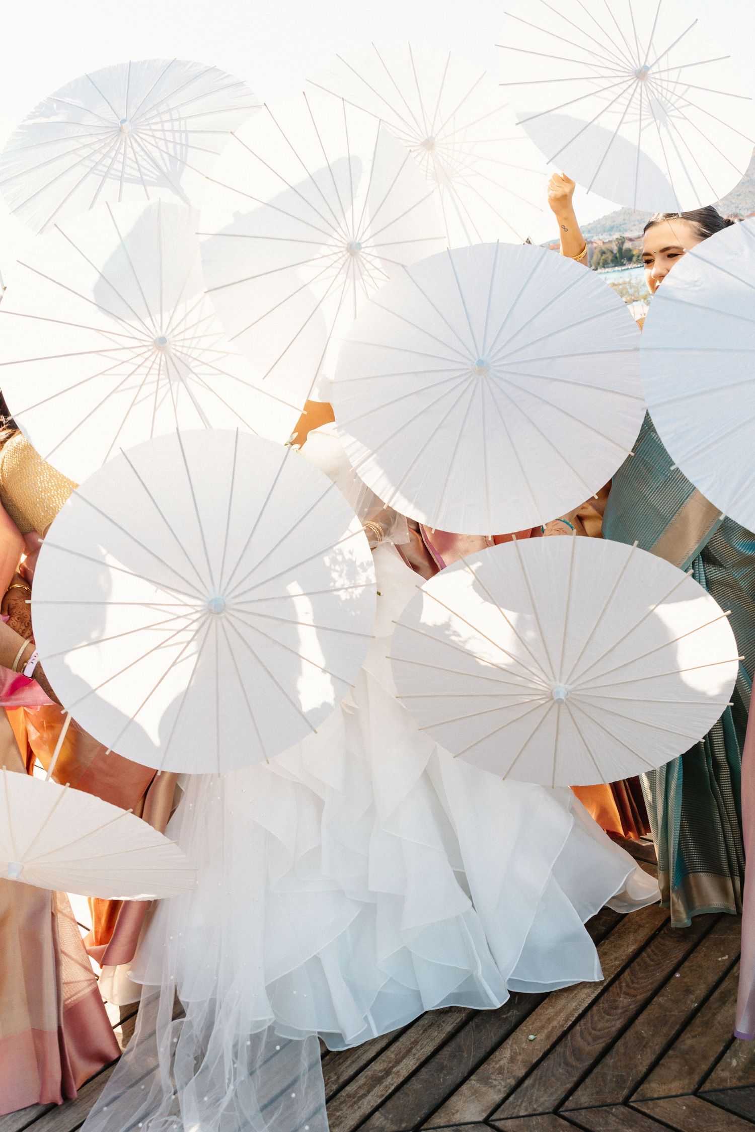 Bride and wedding party holding white parasols creating a stunning canopy of umbrellas on a sunny outdoor deck.