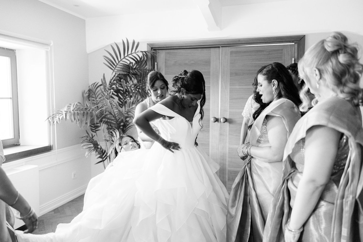 Black and white photo of a bride in a ballgown wedding dress surrounded by bridesmaids in a bright room.
