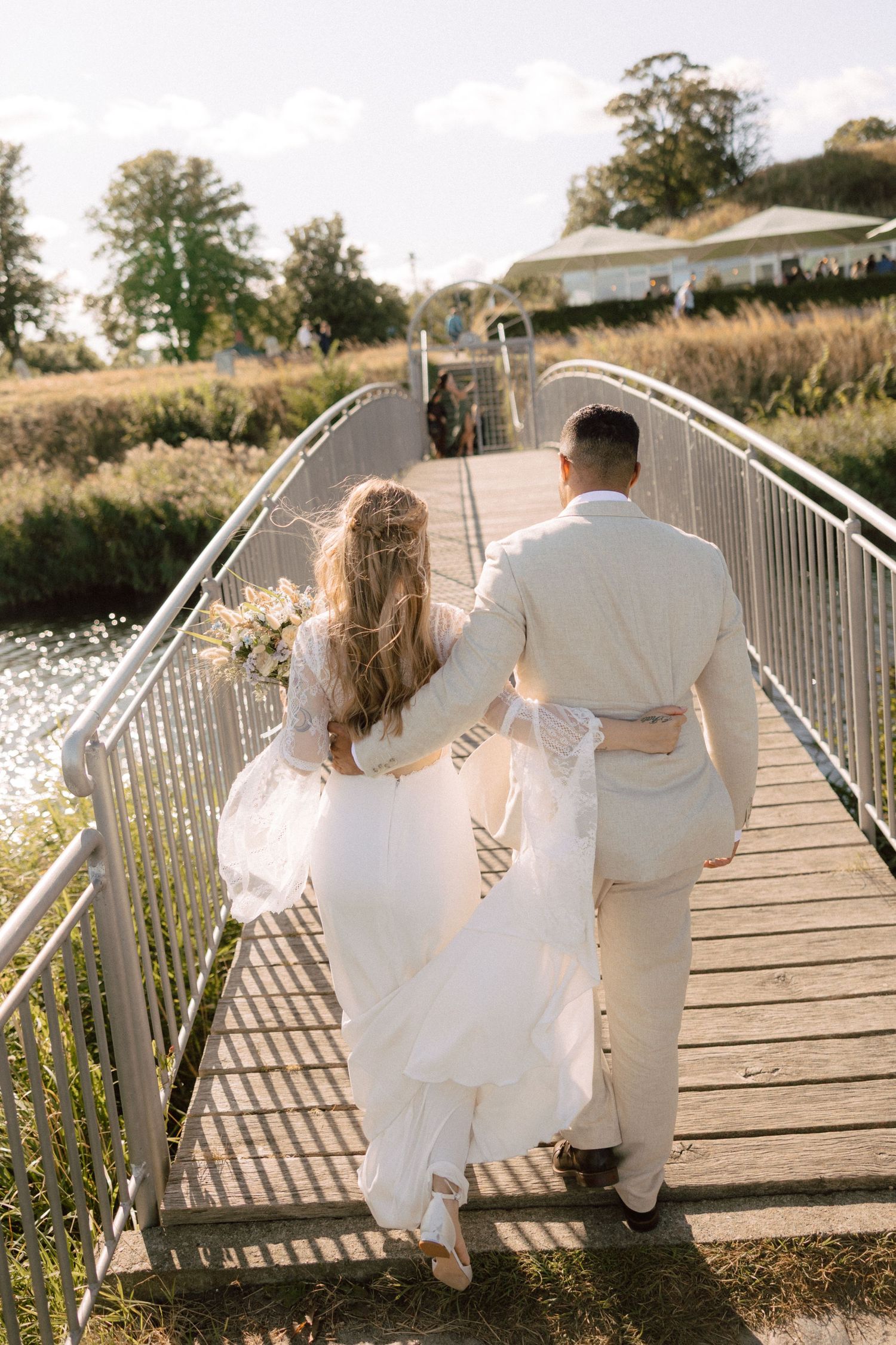 A couple in white wedding attire crosses a wooden bridge surrounded by summer greenery.
