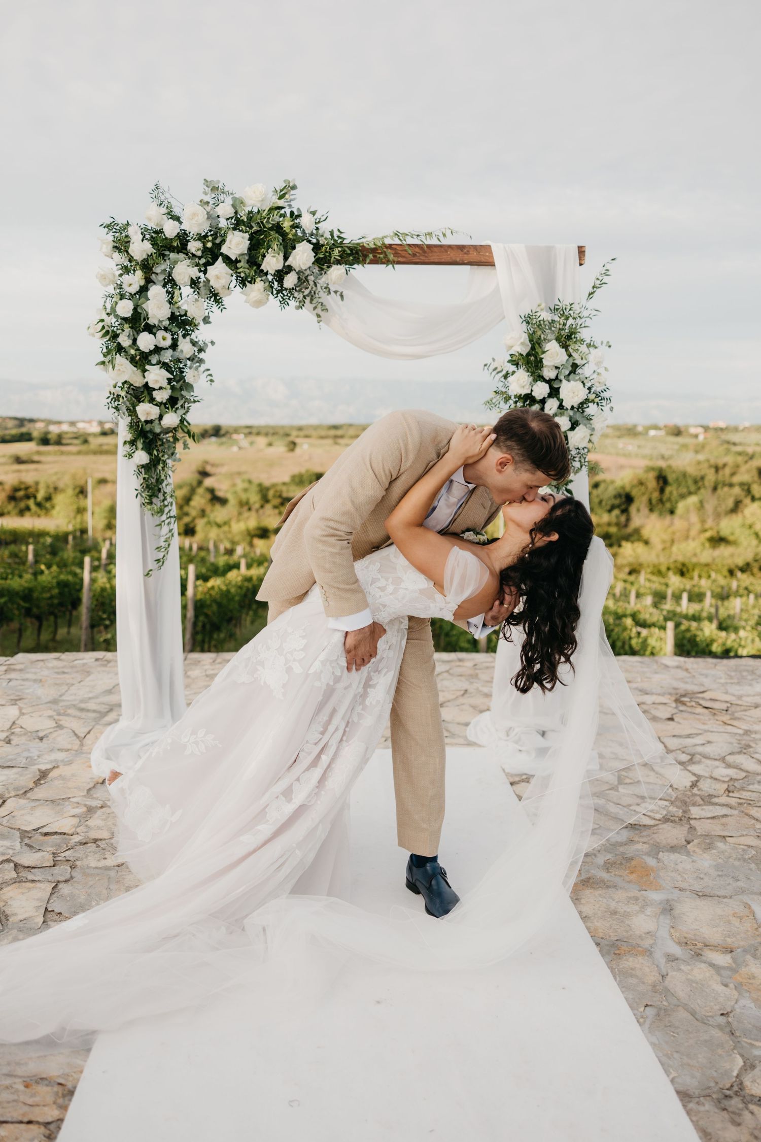 Couple dips and kisses beneath a floral-decorated wedding arch with scenic countryside views.