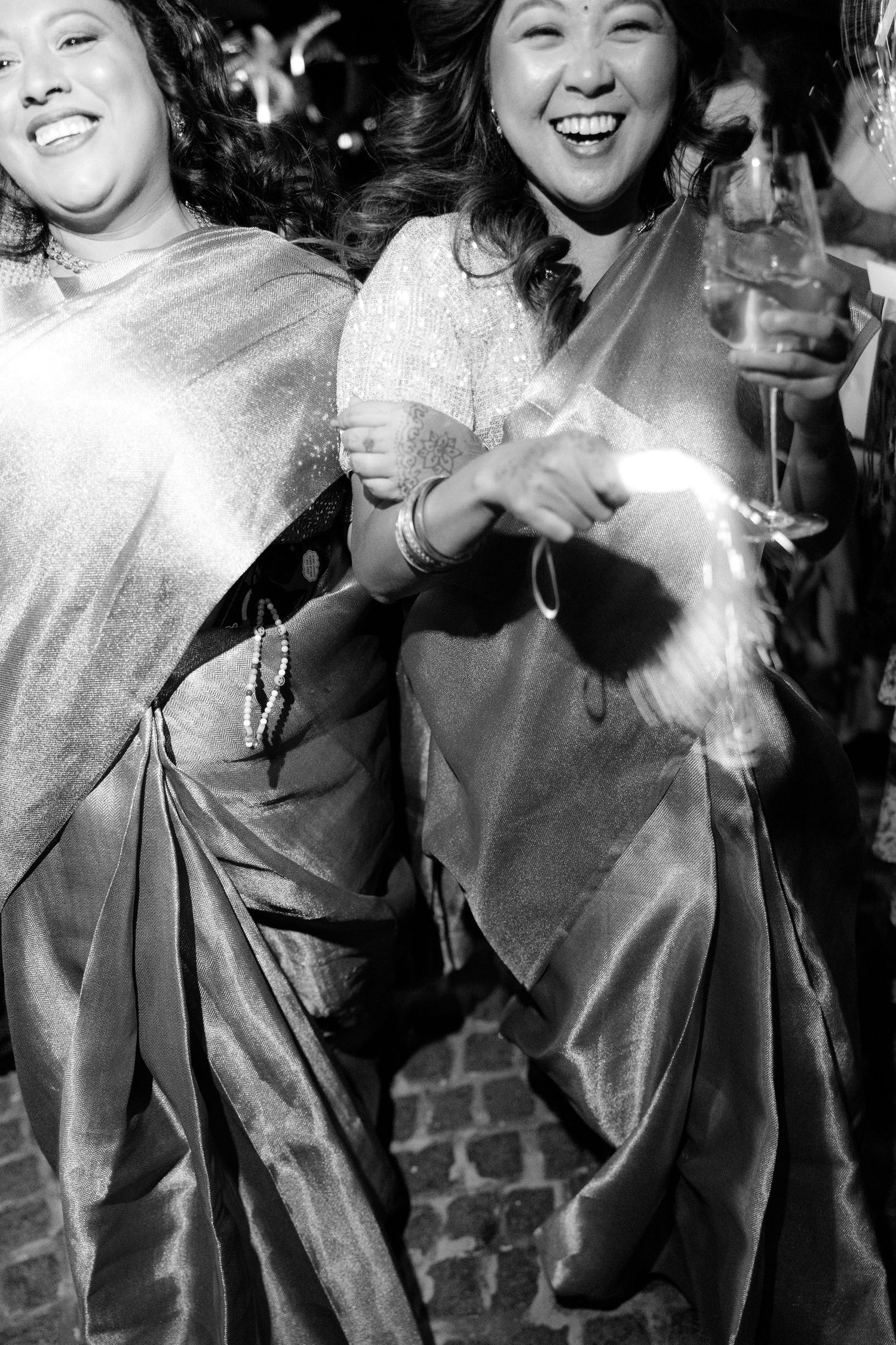 Two women in shimmering saris laugh and dance joyfully at a festive wedding celebration.