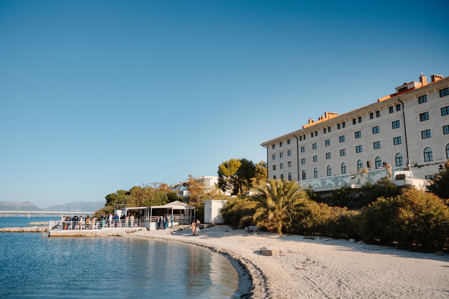 Luxury beachfront hotel with white sandy shore, palm trees, and clear blue sky on a sunny day.