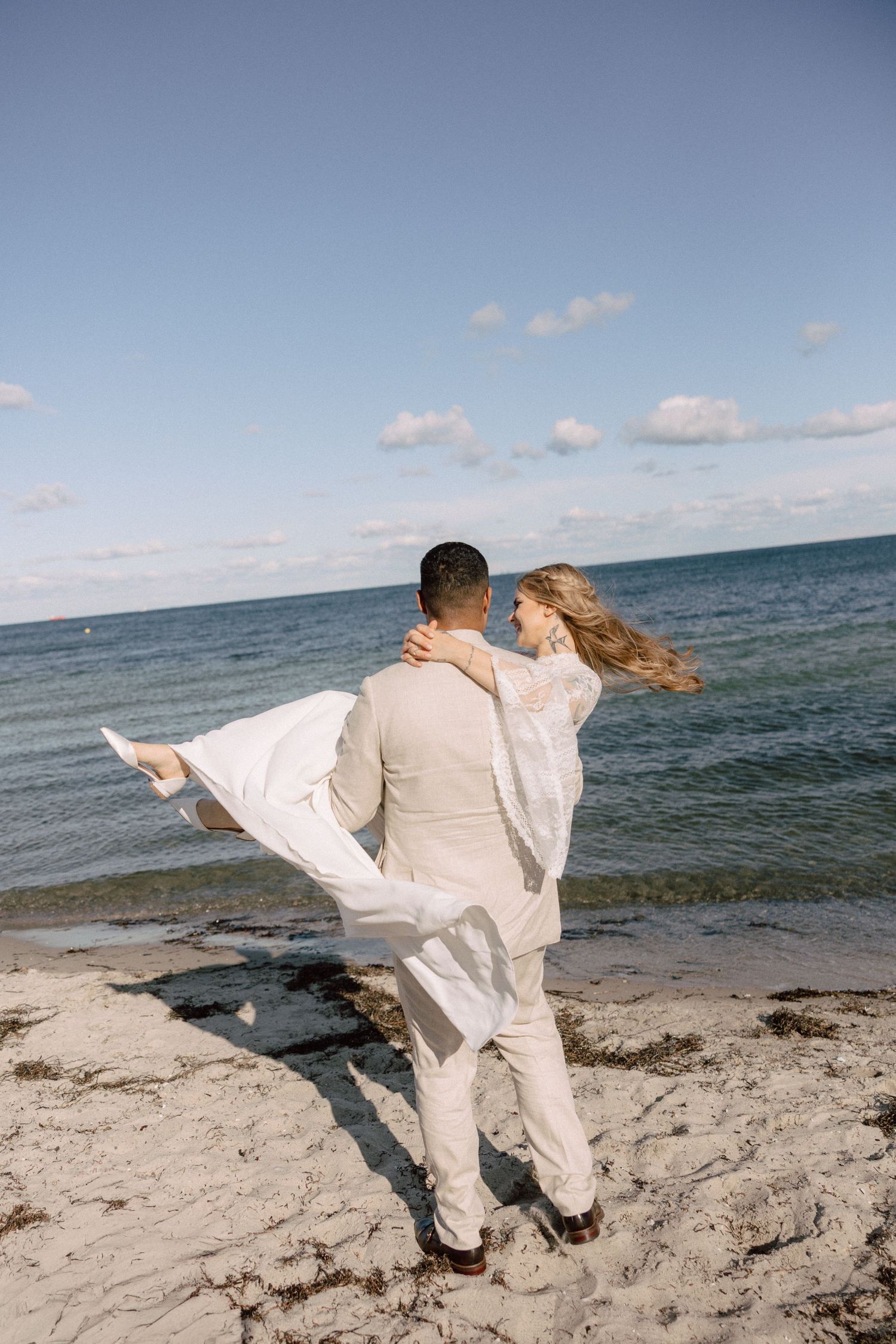 A couple in light-colored wedding attire dances playfully on a beach by the ocean.