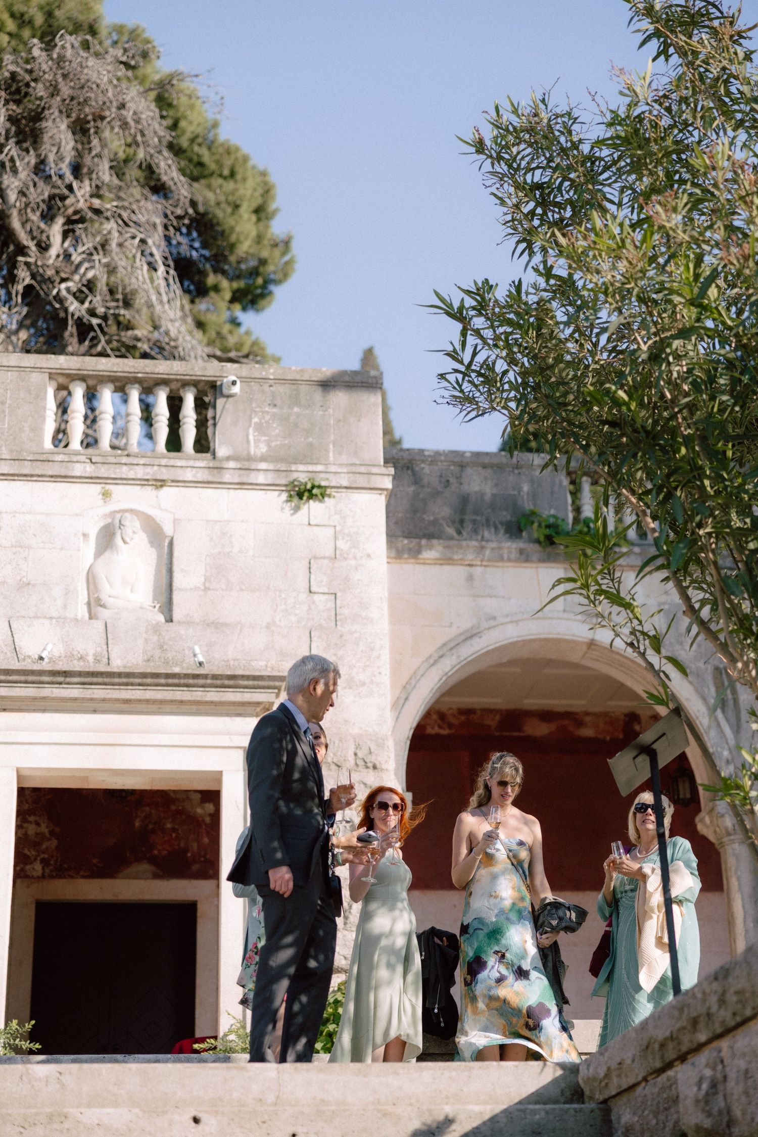 Wedding party gathered on an elegant white stone terrace with arched columns and balustrades overlooking the sea.