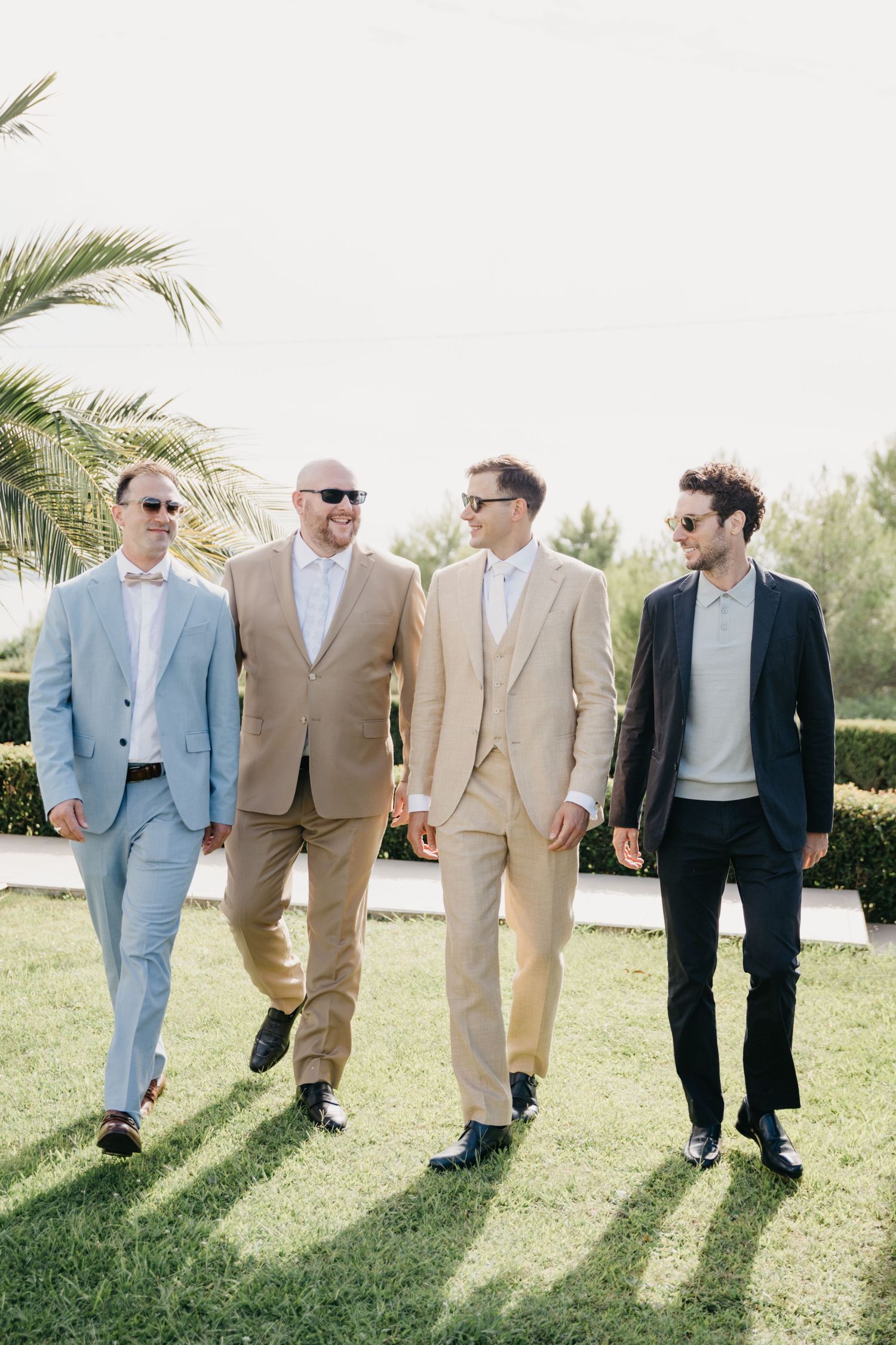 Four groomsmen walking together on grass wearing coordinated suits in light tropical garden setting.