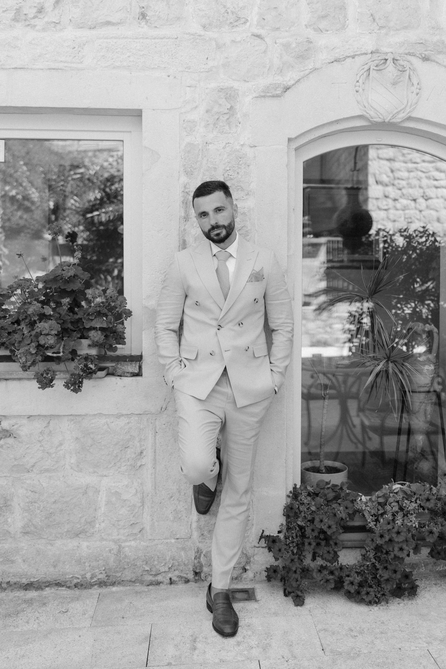 Elegant groom in light suit leaning against stone wall outdoors in black and white portrait photo.