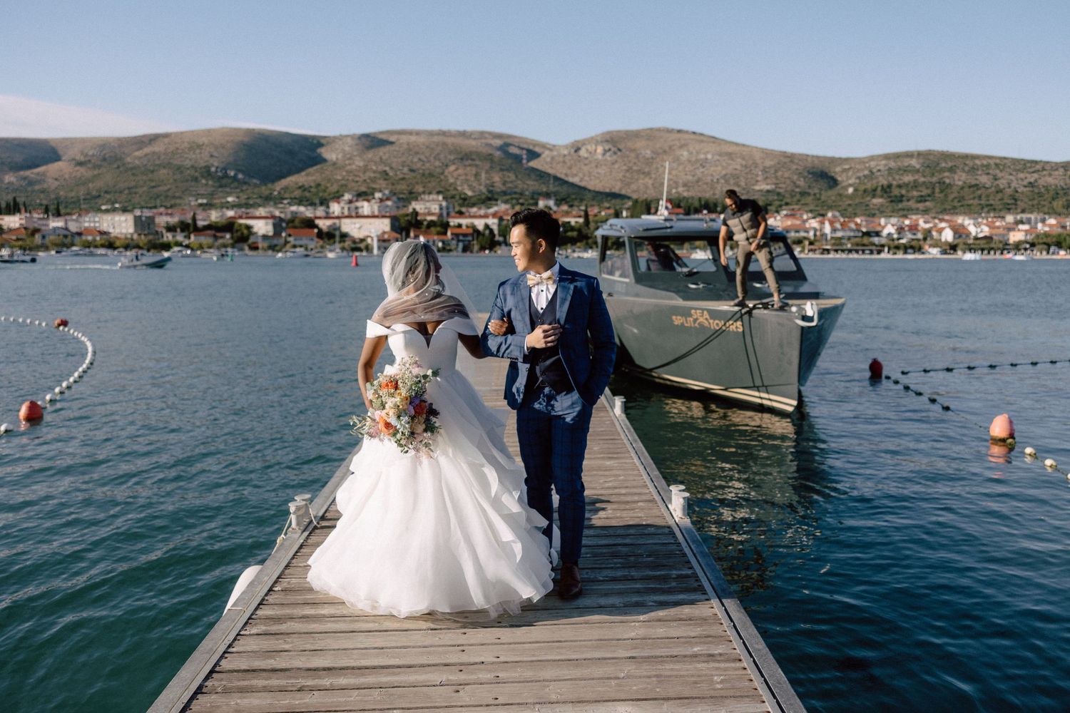 Bride and groom walking hand in hand on dock toward boat on scenic coastal waterway.
