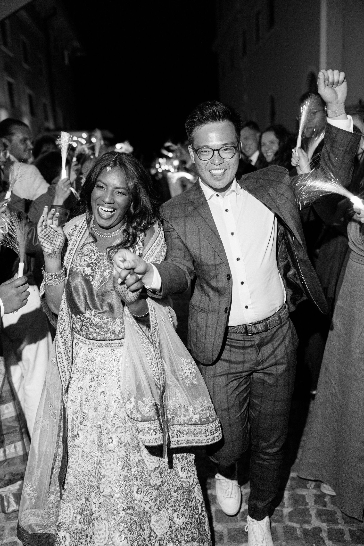 Joyful couple in Indian wedding attire celebrating with guests during festive nighttime baraat procession.