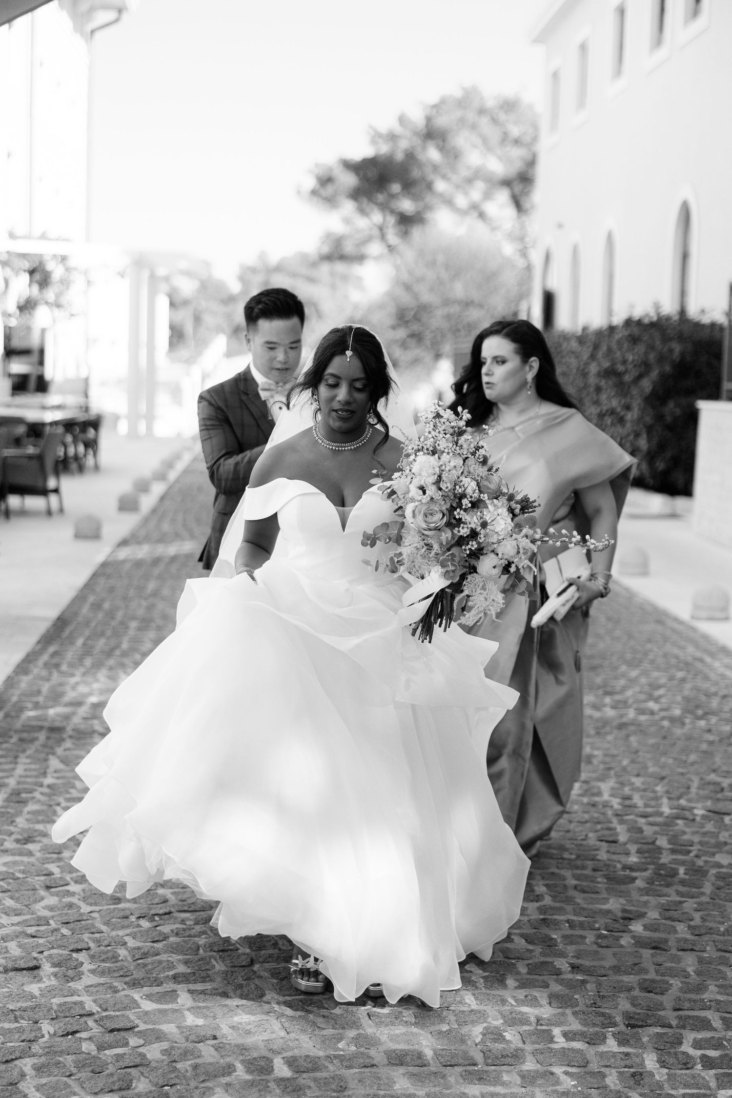 Black and white photo of a bride in a ball gown walking on cobblestones assisted by two wedding guests.