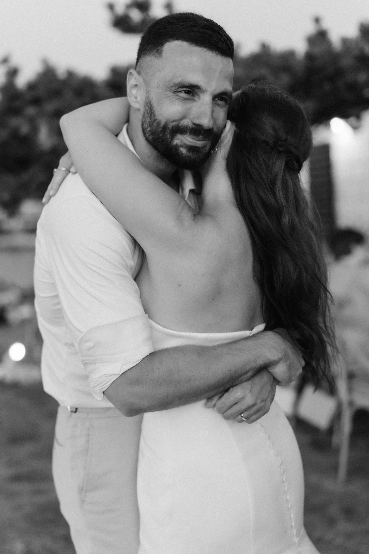 Black and white portrait of smiling groom embracing bride in a backless wedding dress at dusk.