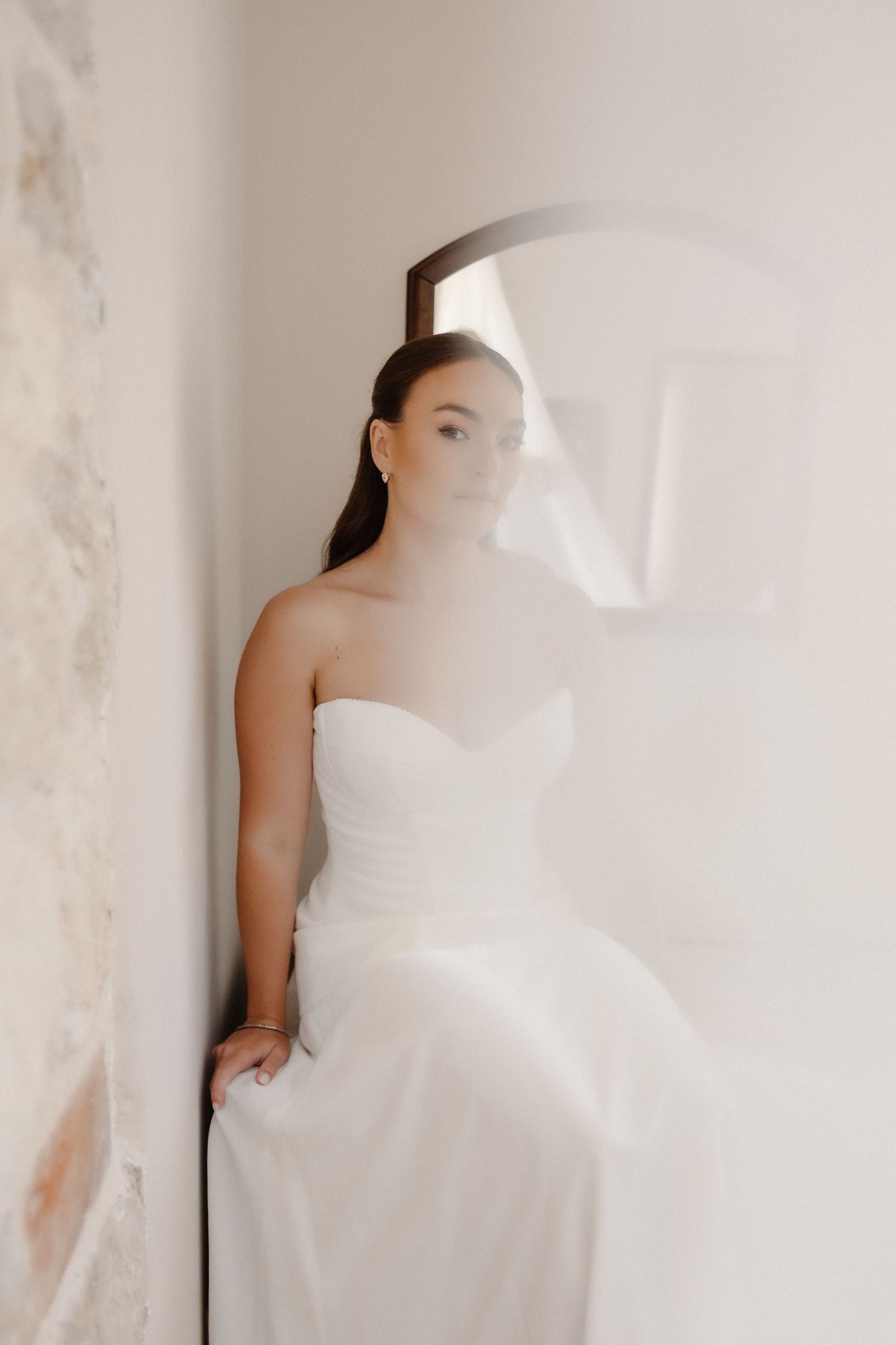 Bride in a strapless white gown poses gracefully by an arched window, her veil softly blurring in the light.