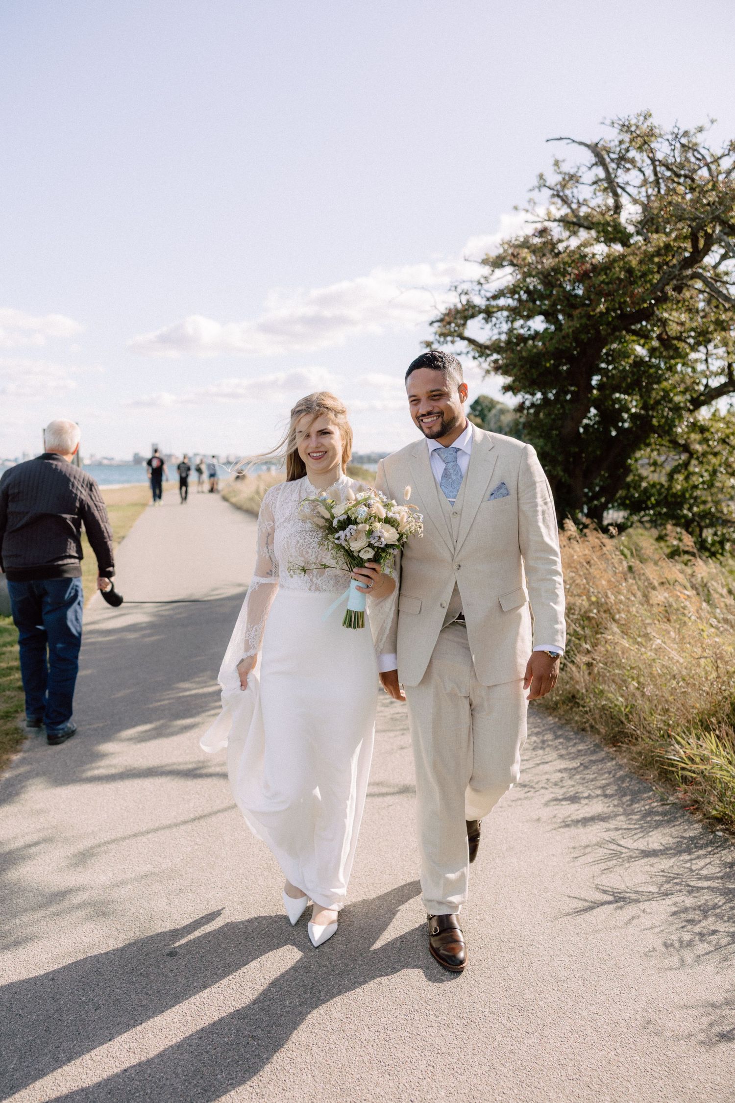 A newlywed couple walks along a seaside path in elegant white and beige wedding attire.