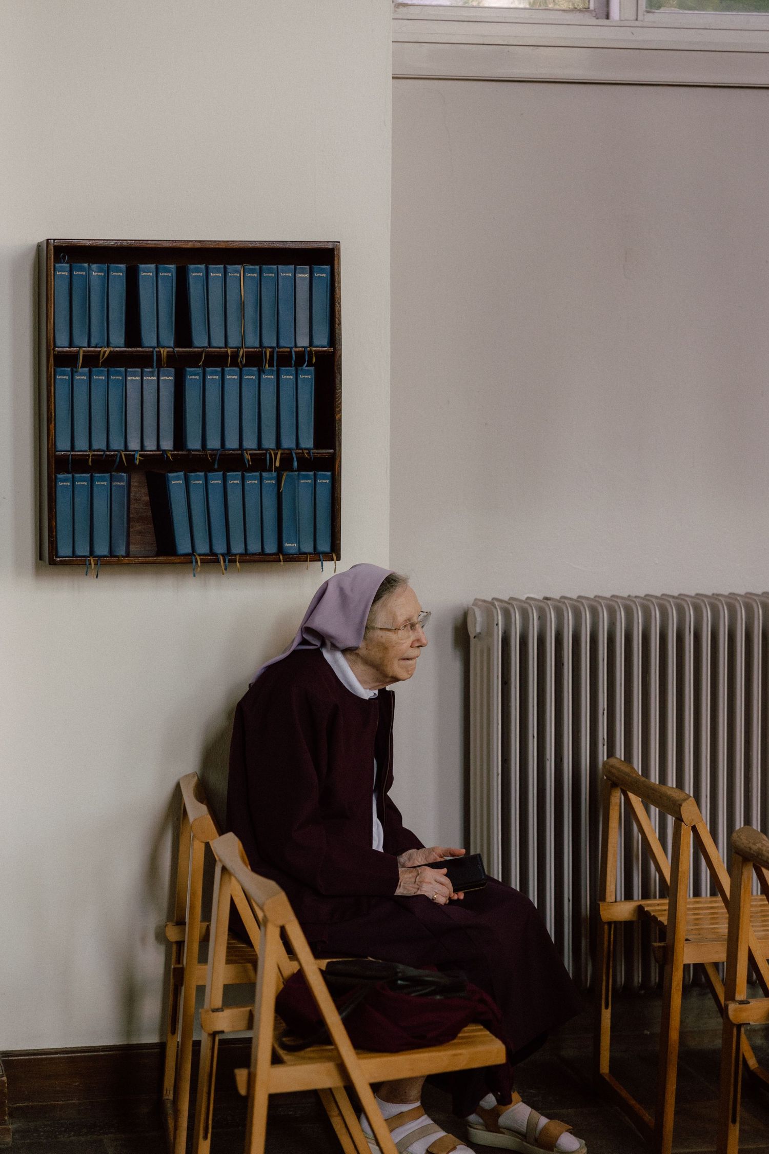 Person sits alone on wooden chair against white wall with blue books on shelf above.