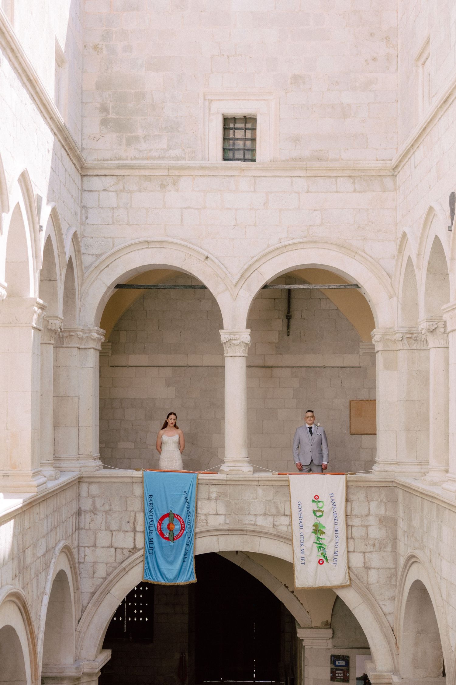 Wedding couple photographed in a grand monastery courtyard with hanging banners and stone arches in Dubrovnik, Croatia.