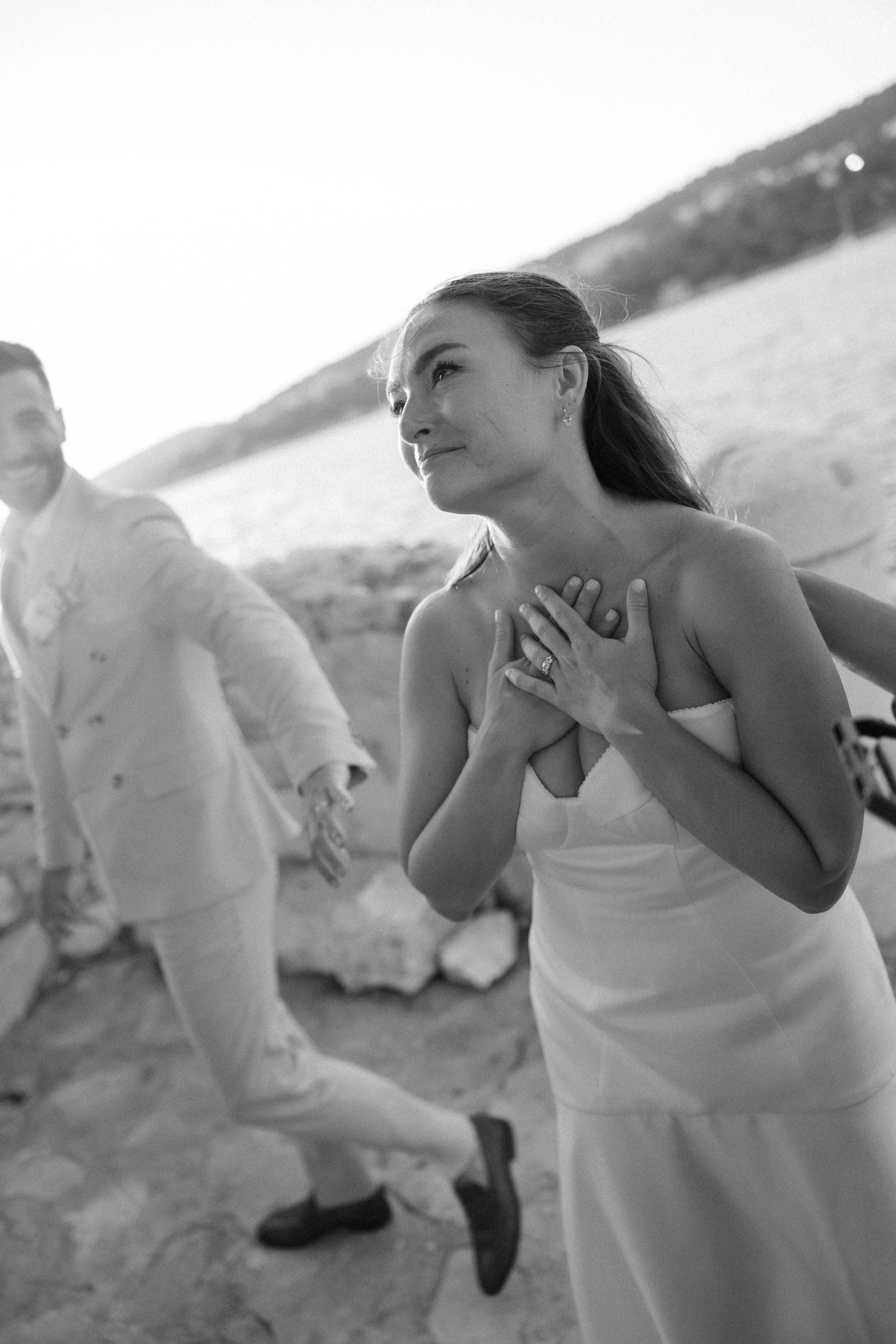 Emotional bride in white dress holds hands to chest during outdoor beach wedding ceremony.