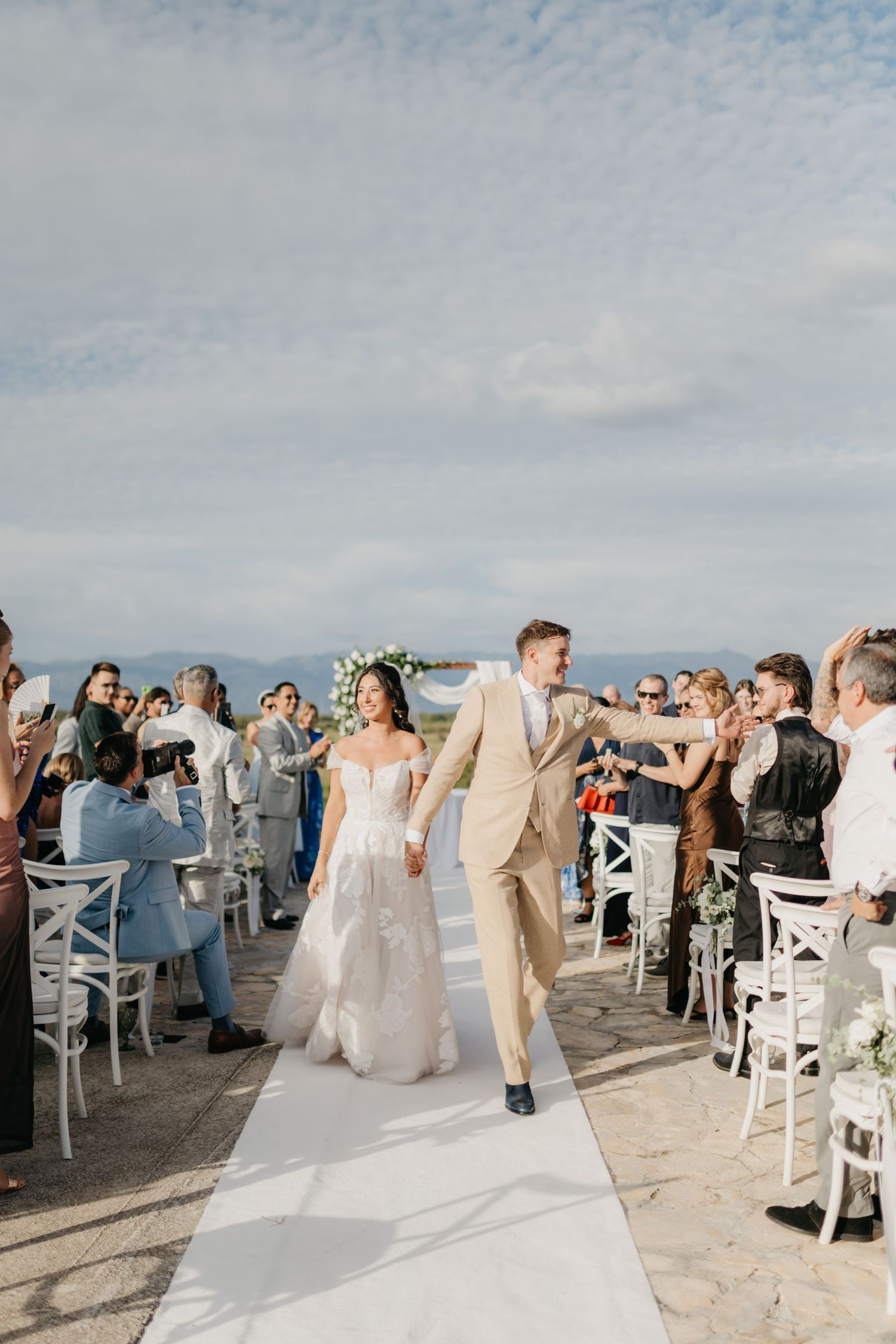 Wedding guests stand and cheer as the newlyweds make their entrance at an outdoor venue in Italy.