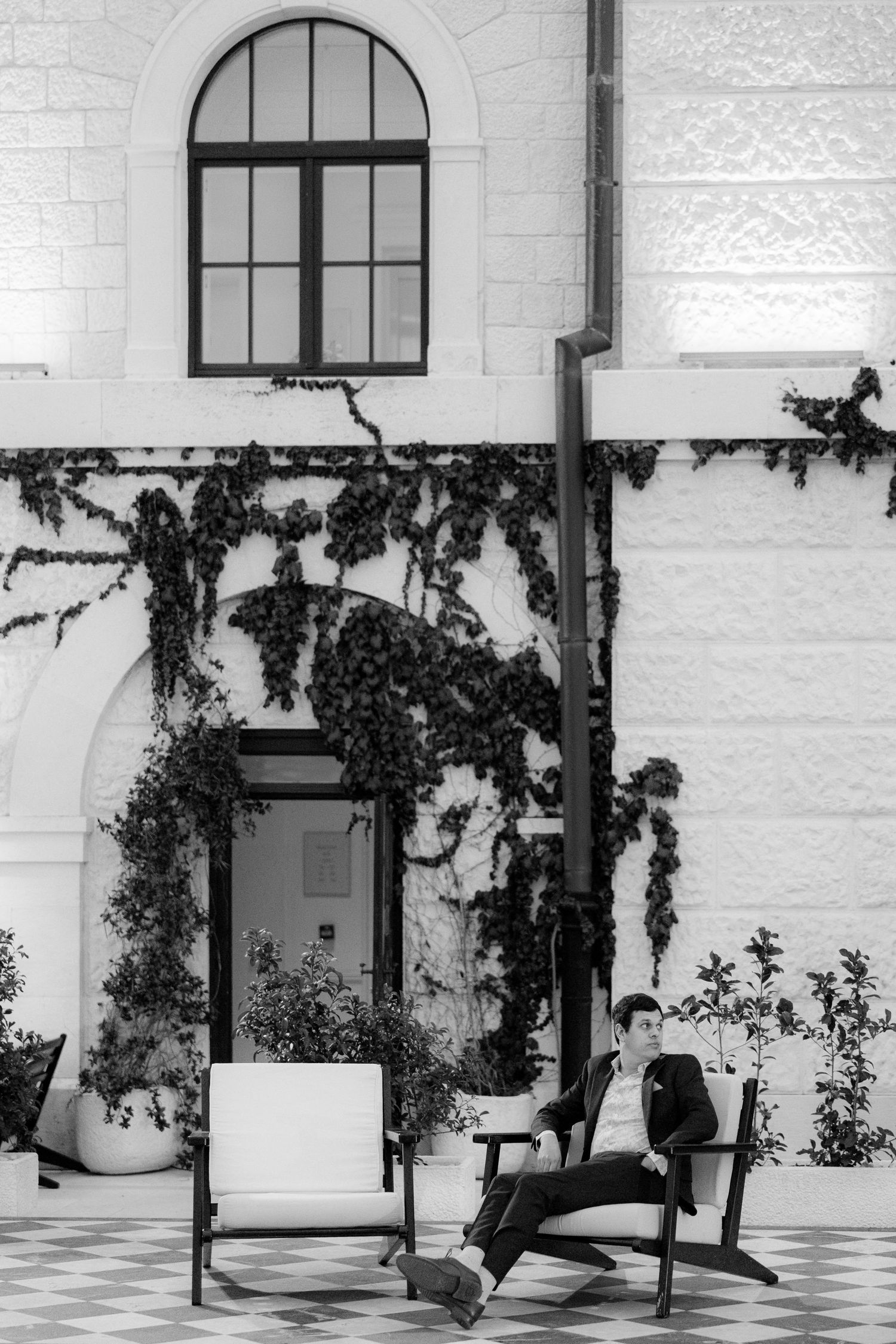 Groom relaxes in chair beside ivy-covered white building wall in black and white portrait.