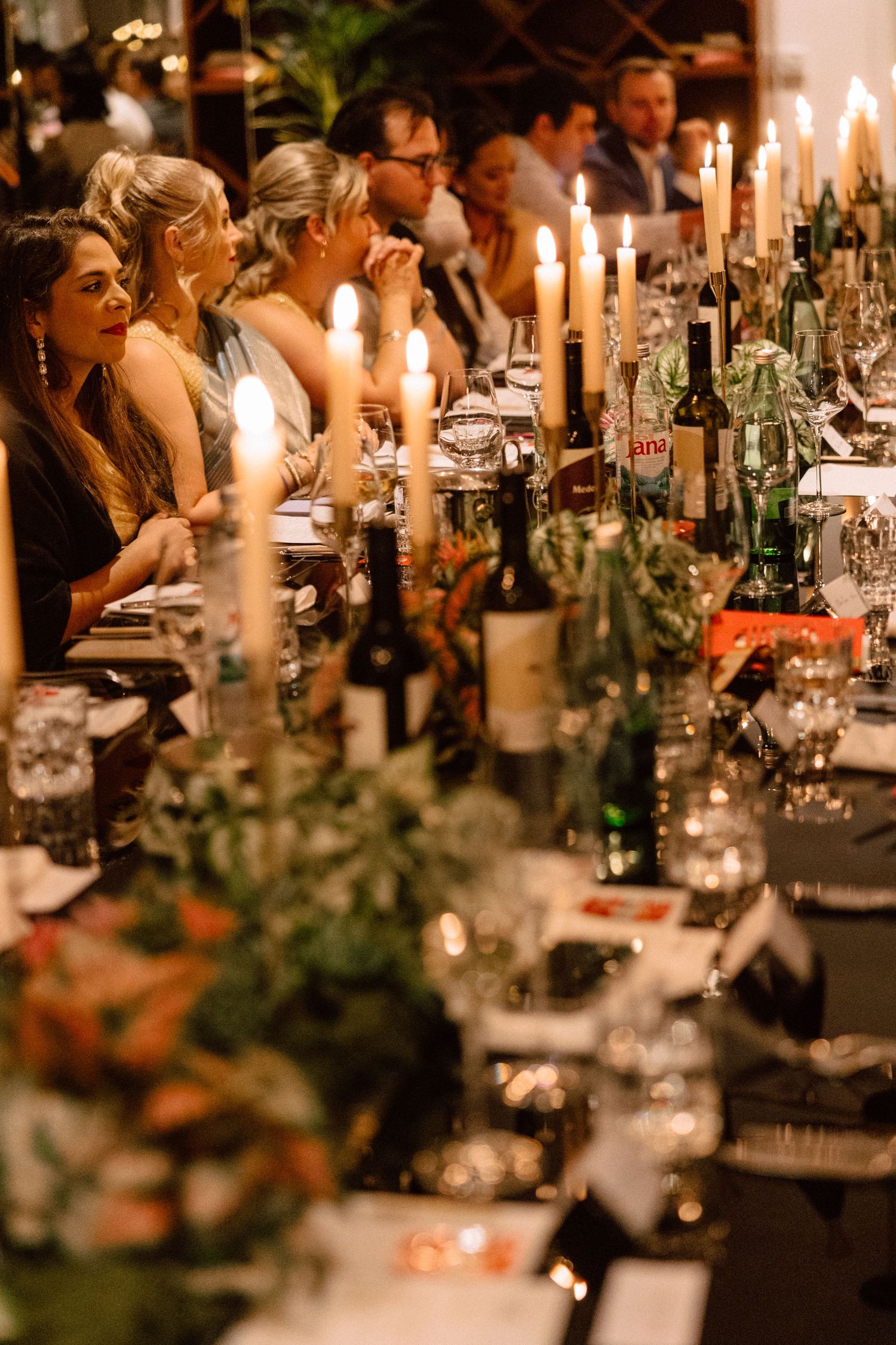 Elegant candlelit wedding reception dinner table adorned with greenery, wine bottles, and glassware with guests seated alongside.