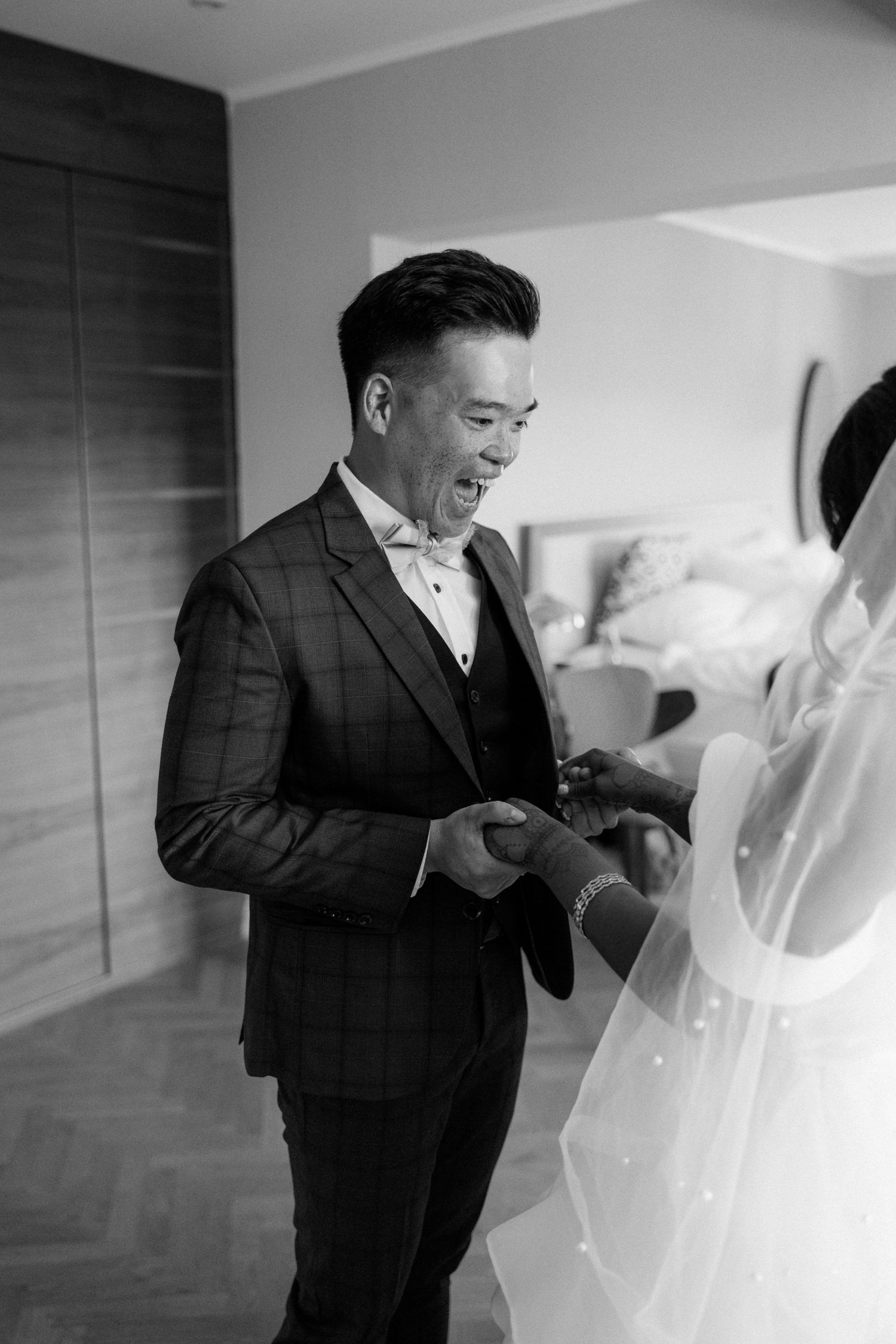 Black and white photo of a smiling Asian groom in a plaid suit holding hands with his veiled bride.