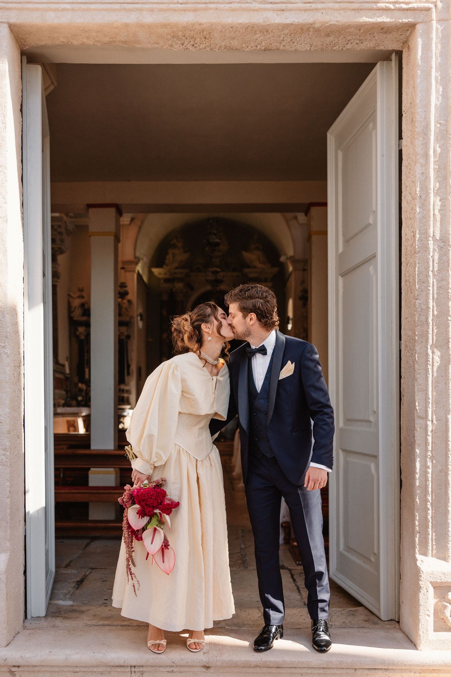 A romantic wedding couple sharing a kiss in a doorway while holding a red bouquet.