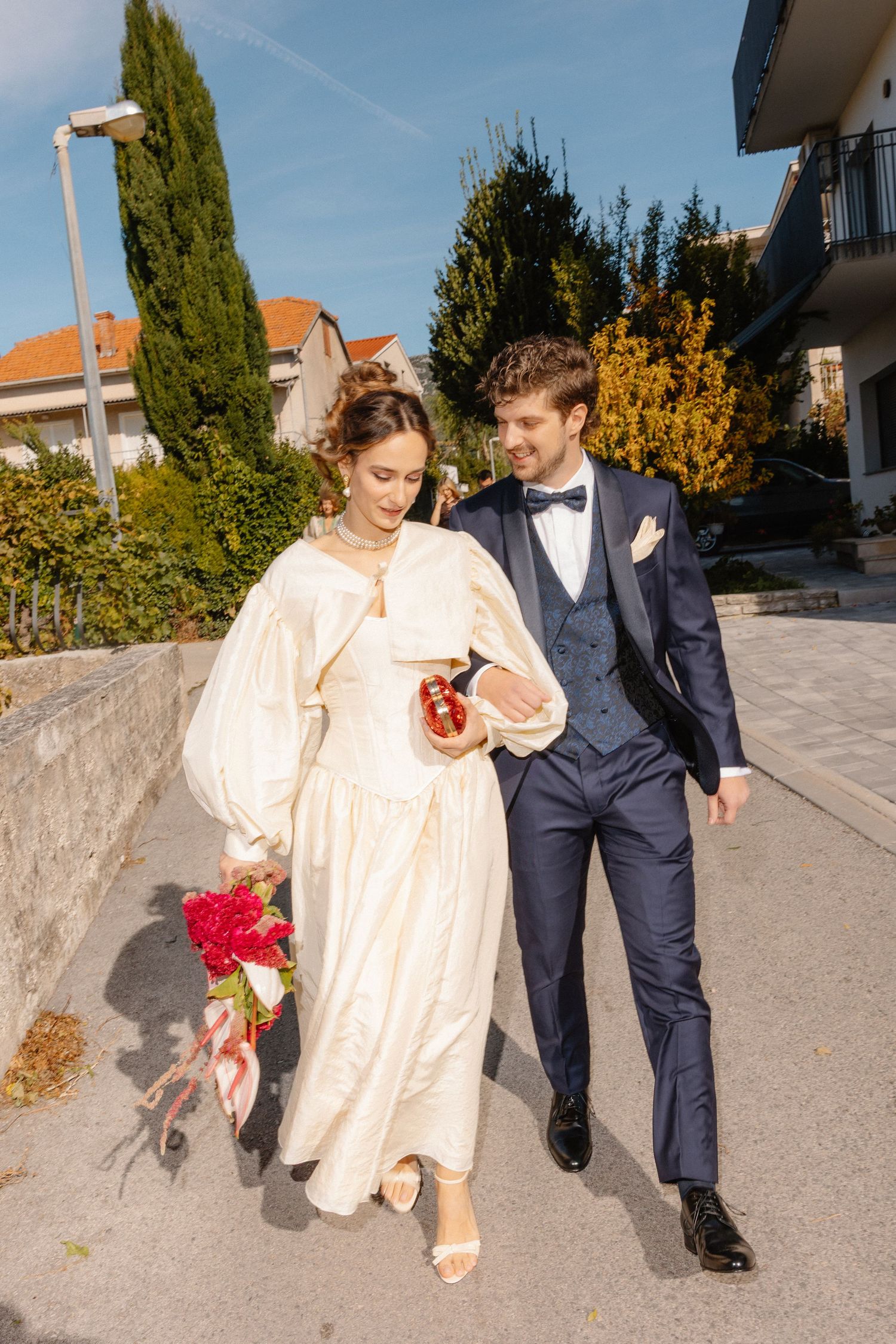 A couple walking together outdoors, with the person in cream-colored dress holding red flowers and navy suit companion.