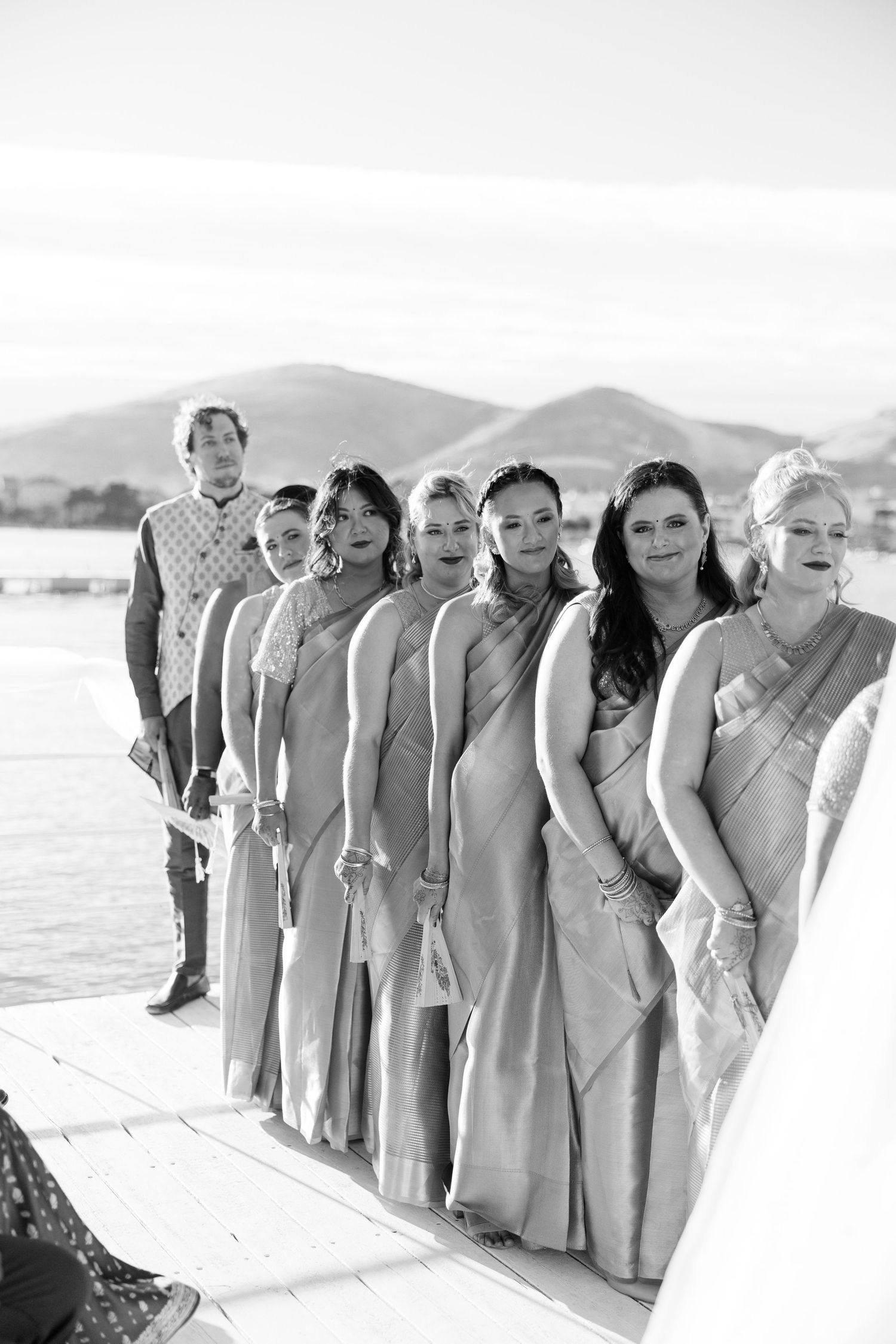 Black and white photo of bridesmaids in matching sarees standing in a line outdoors near water with mountains in background.