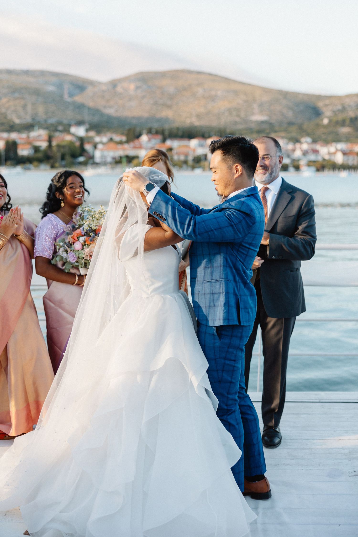 Groom in blue suit lifting bride's veil during outdoor waterfront wedding ceremony with guests watching.