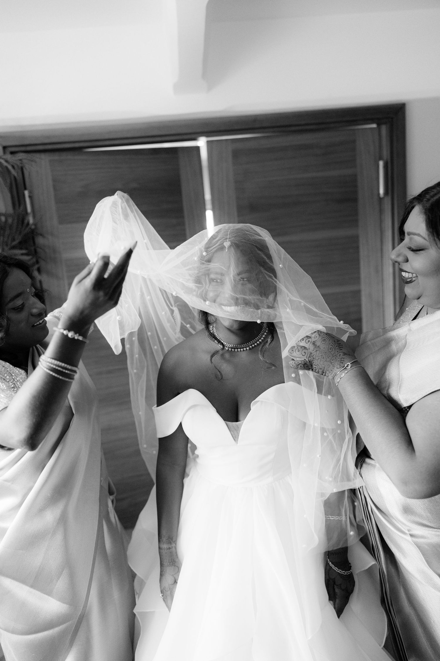 Black and white photo of bridesmaids placing a pearl-trimmed veil over a smiling bride in white gown.
