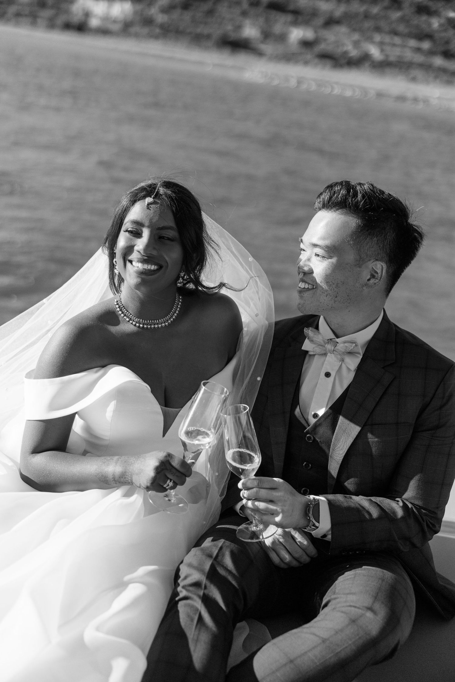 Black and white photo of a smiling interracial couple toasting with champagne glasses by the water on their wedding day.