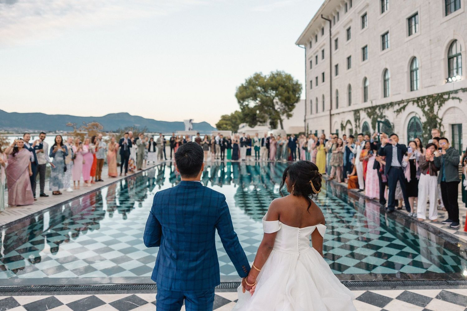 Bride and groom walk hand-in-hand toward cheering guests by a reflective pool.