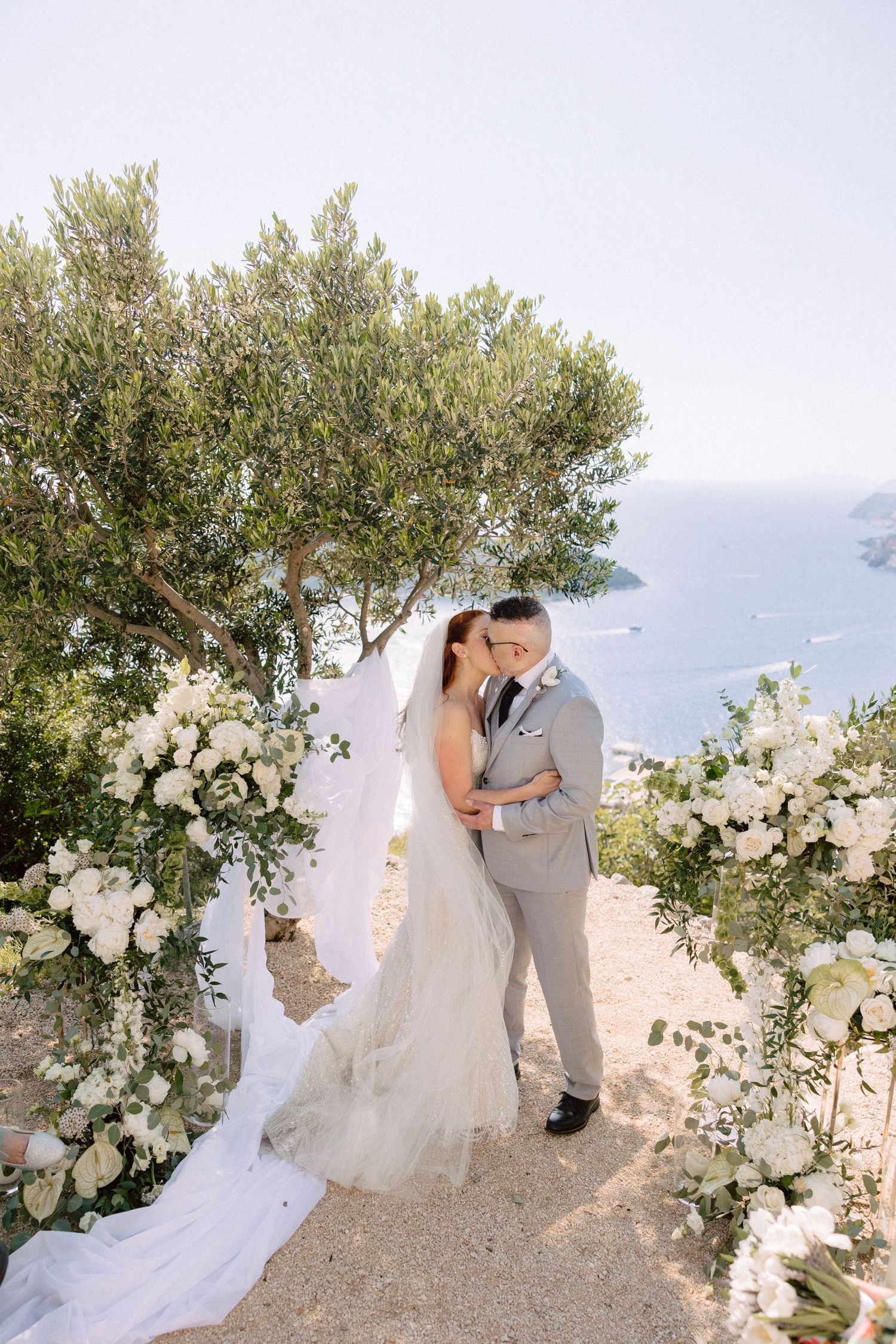 A wedding couple shares intimate moments under an olive tree arch decorated with white flowers.