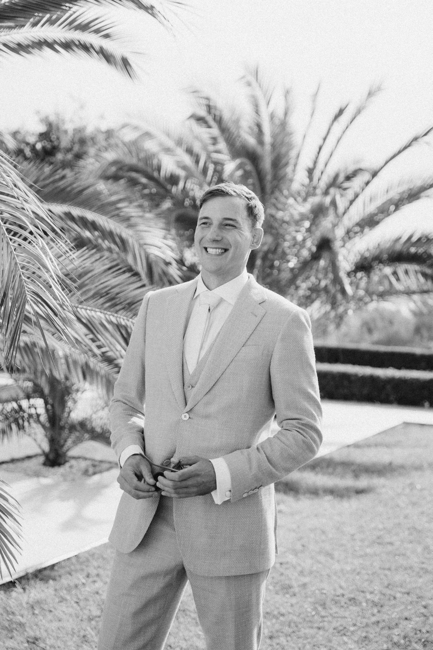 A groom in a light gray suit smiles while standing in front of palm trees on his wedding day.