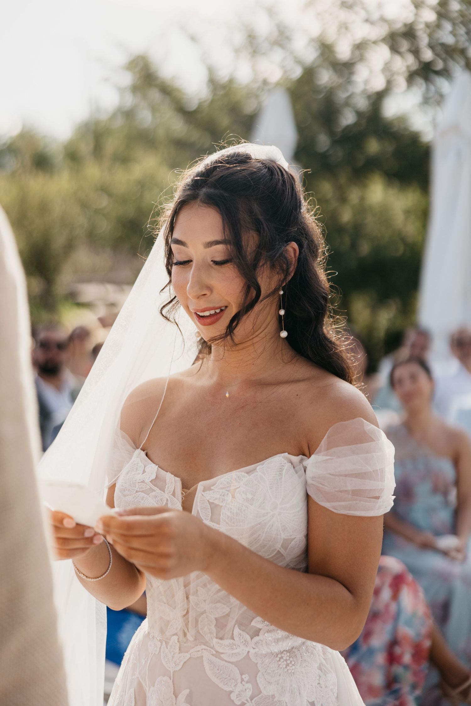 A bride in a white off-shoulder dress reads from a paper during an emotional outdoor ceremony.