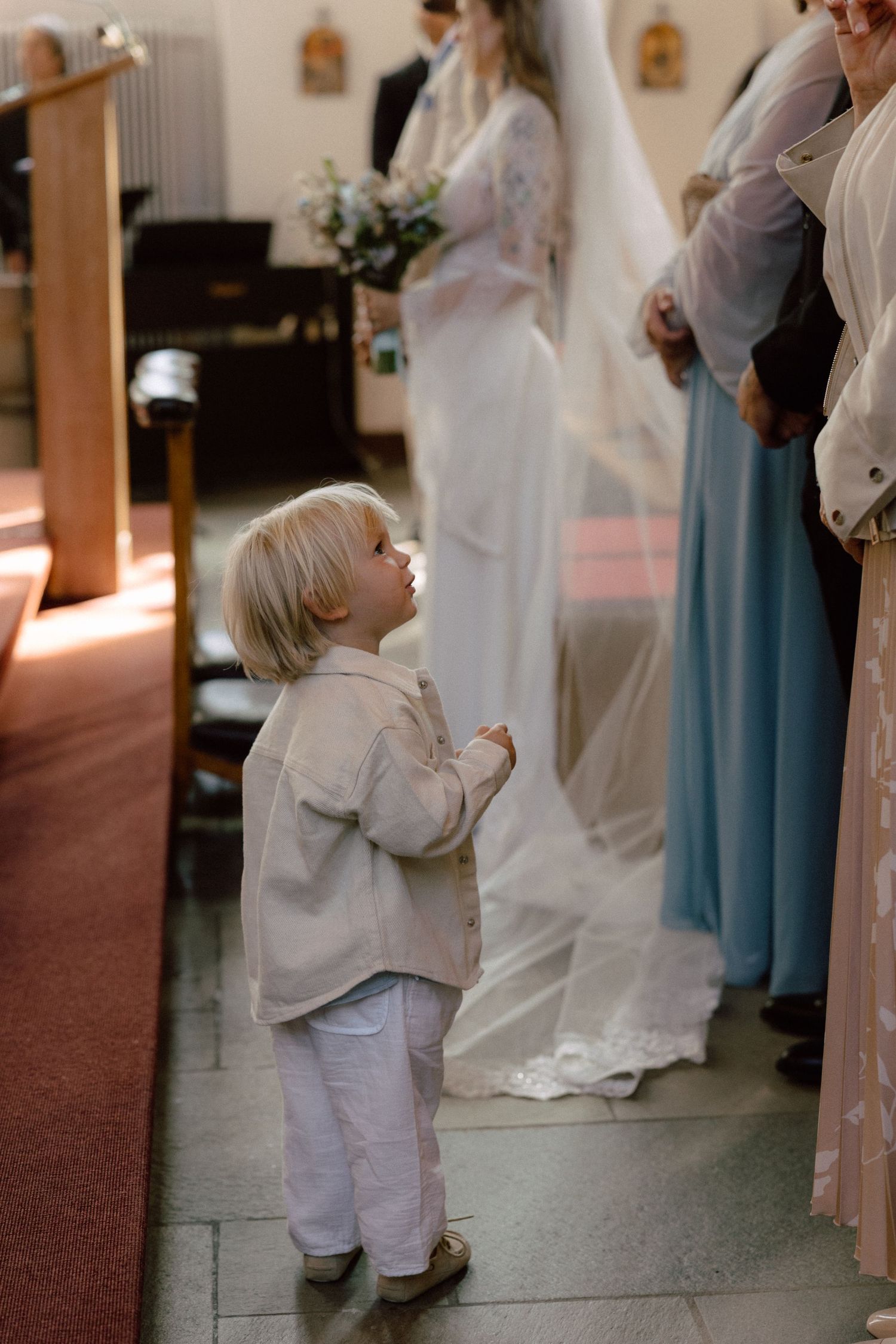 Small child in white outfit stands near wedding party during church ceremony.