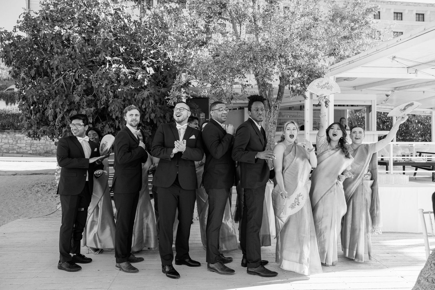 Wedding party posing playfully in black and white, bridesmaids in saris and groomsmen in suits outdoors.