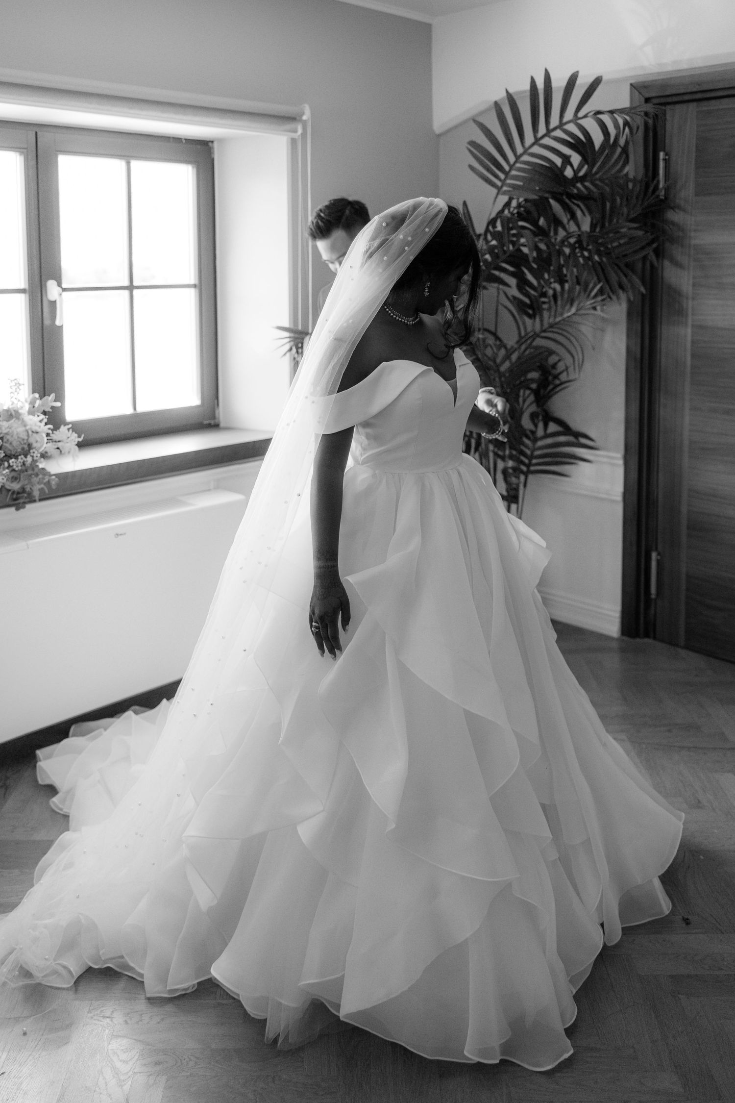 Elegant bride in a ruffled off-shoulder wedding gown with veil poses by window in black and white photo.