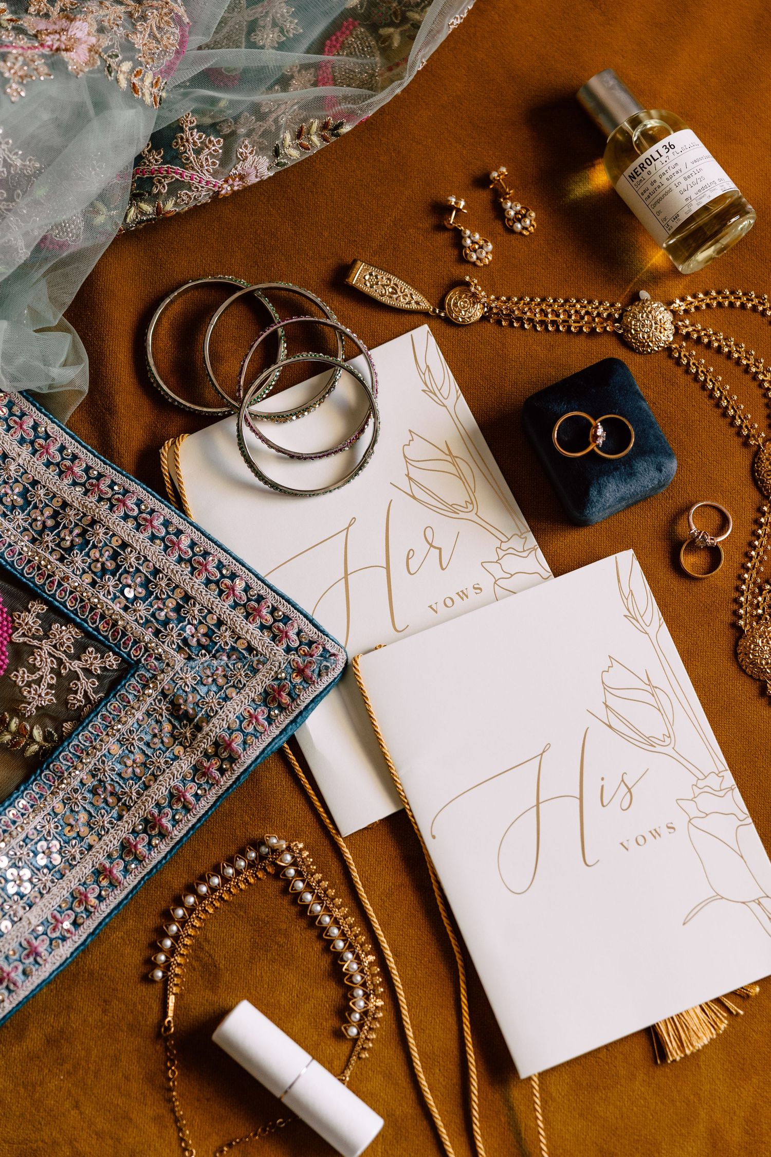 Elegant wedding flat lay with vow books, gold jewelry, bangles, rings, and perfume on a warm brown surface.