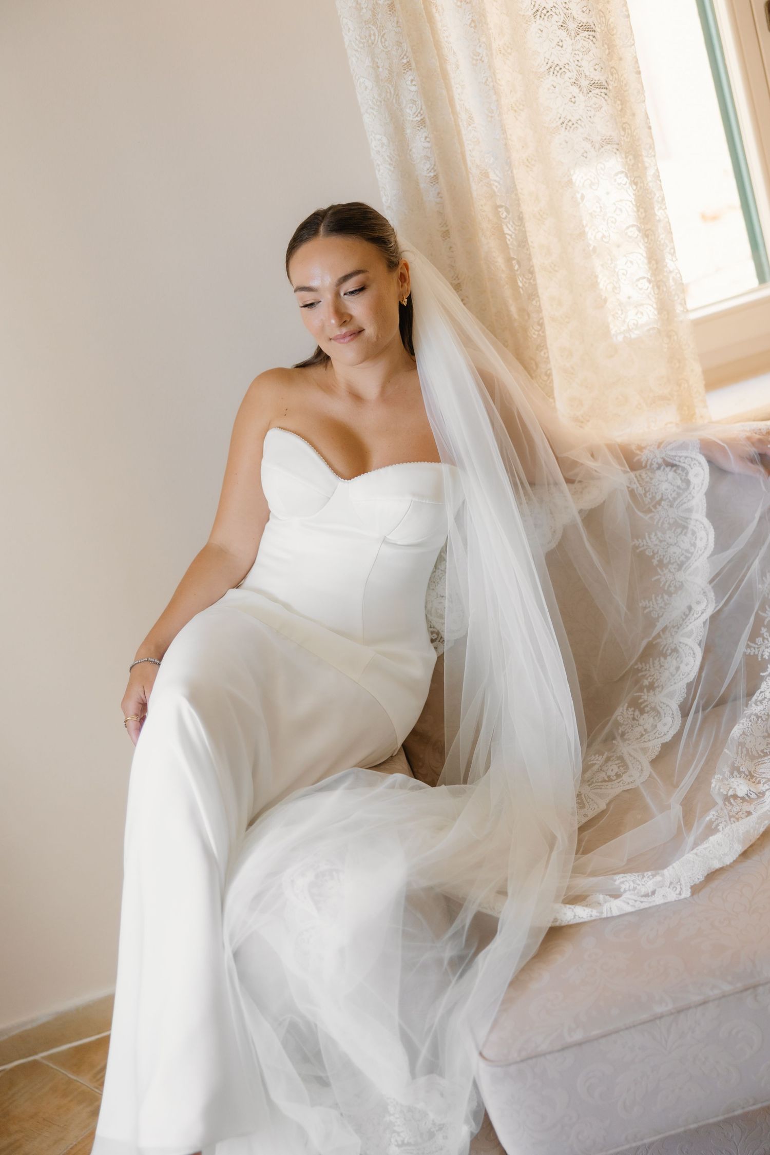 Bride in strapless white jumpsuit with flowing veil seated by window in soft natural light.