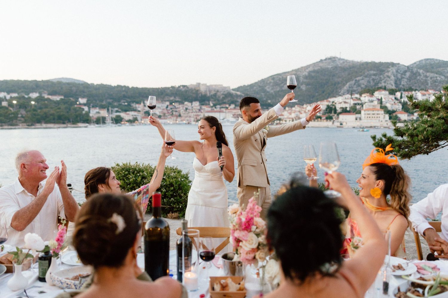 Bride and groom toast with guests at outdoor waterfront wedding reception with scenic harbor views.
