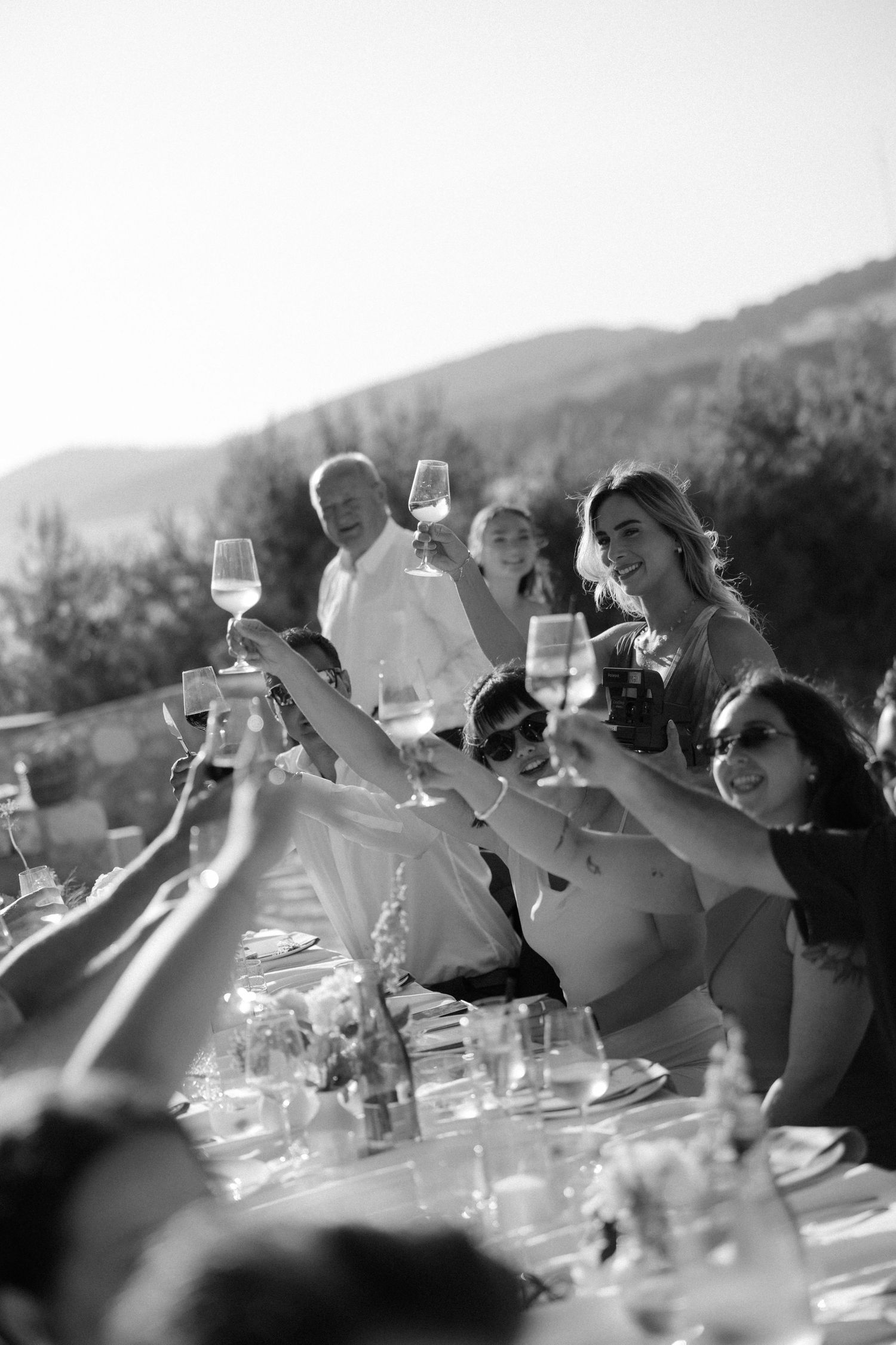 Black and white photo of wedding guests raising champagne glasses in joyful outdoor toast.