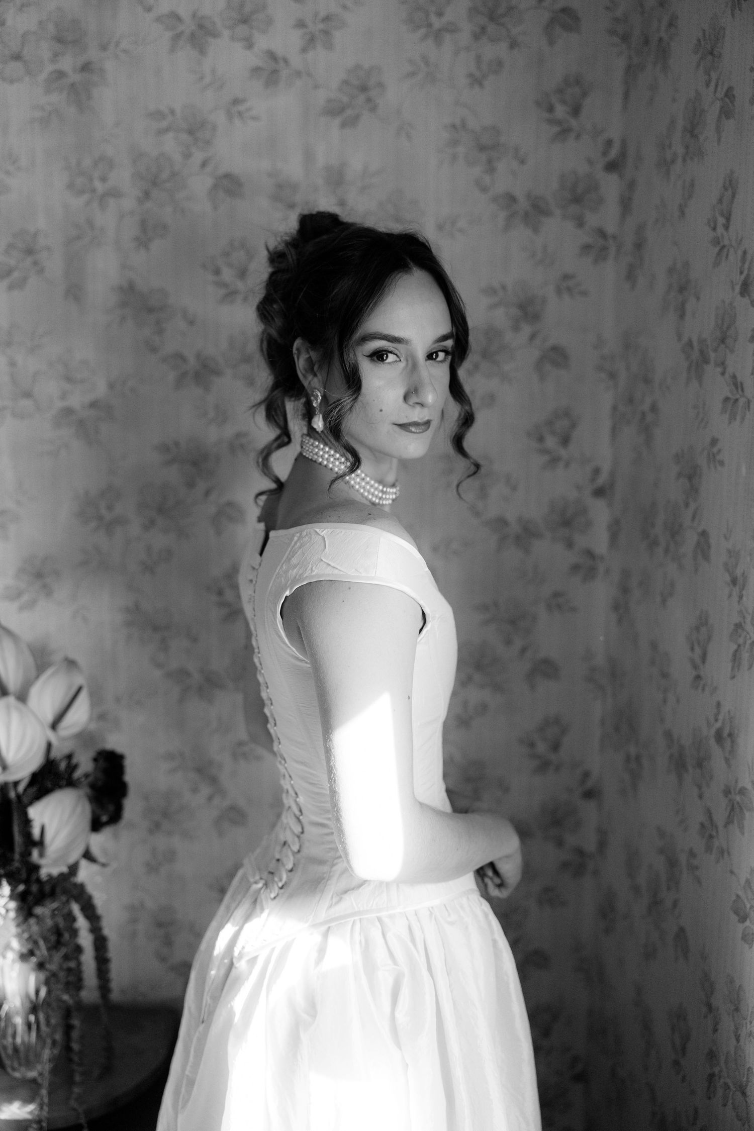 A black and white portrait series showing someone in a white Victorian-style dress against a patterned wallpaper background.