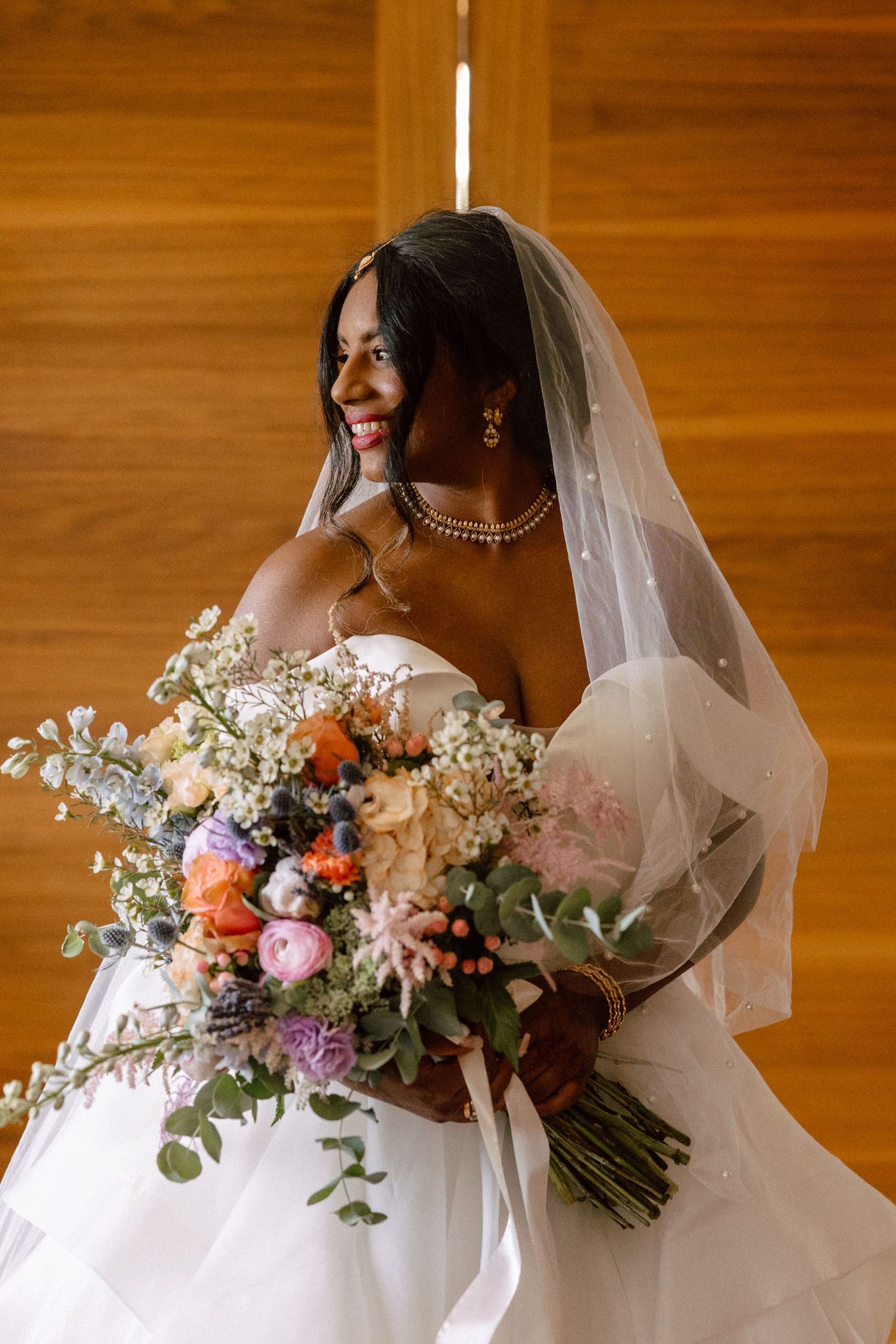 Smiling bride in white gown and veil holding vibrant mixed floral bouquet in wooden chapel.