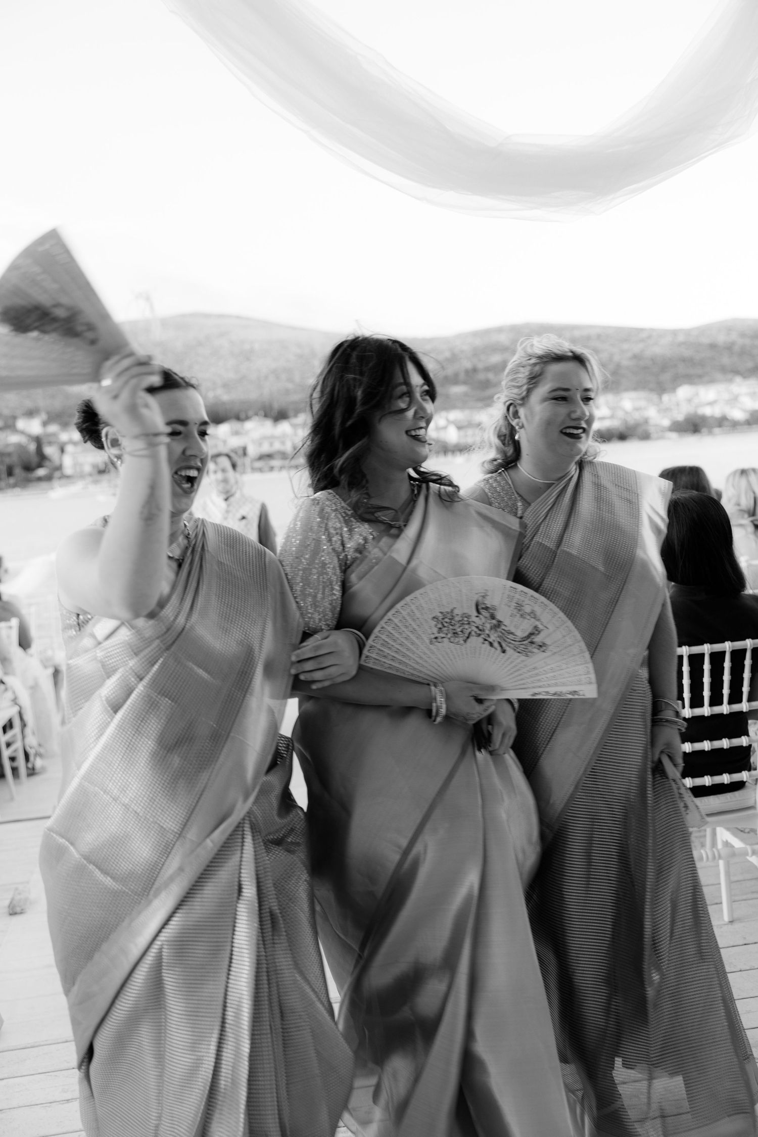 Three bridesmaids in silk sarees laugh joyfully holding fans at outdoor wedding ceremony.