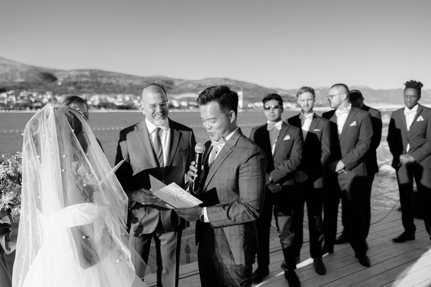 Groom reads vows at waterfront wedding ceremony with groomsmen standing beside him in black and white.