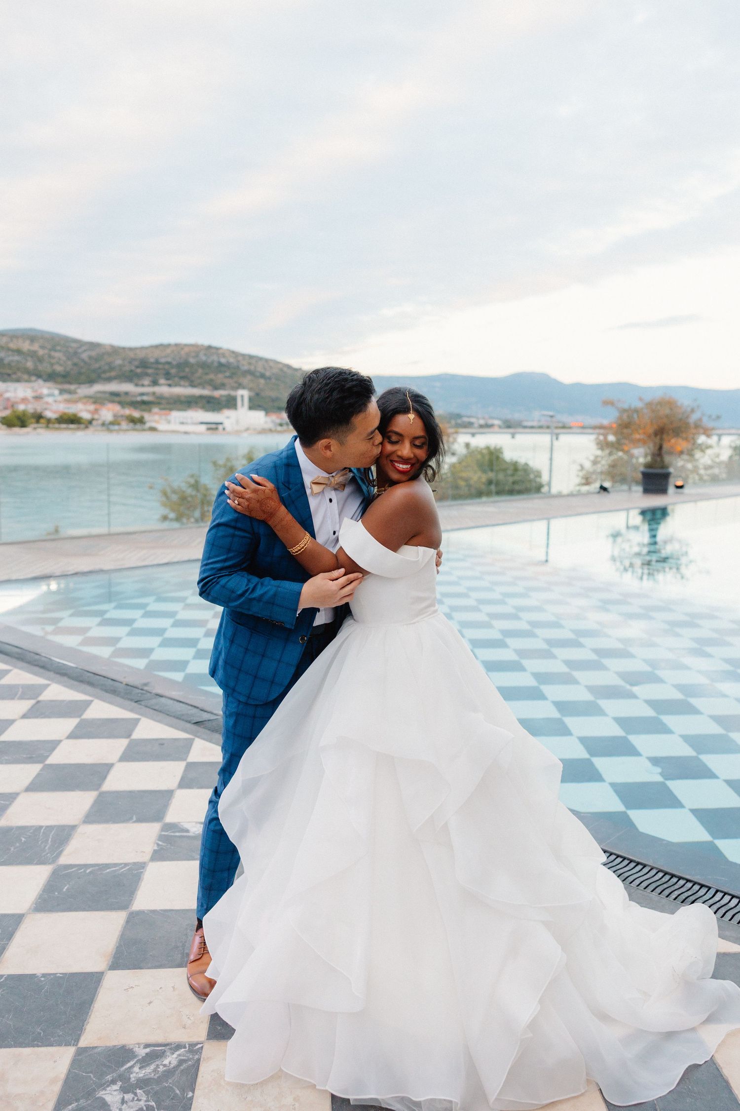 Groom in blue suit embracing bride in layered white off-shoulder gown on scenic waterfront terrace.