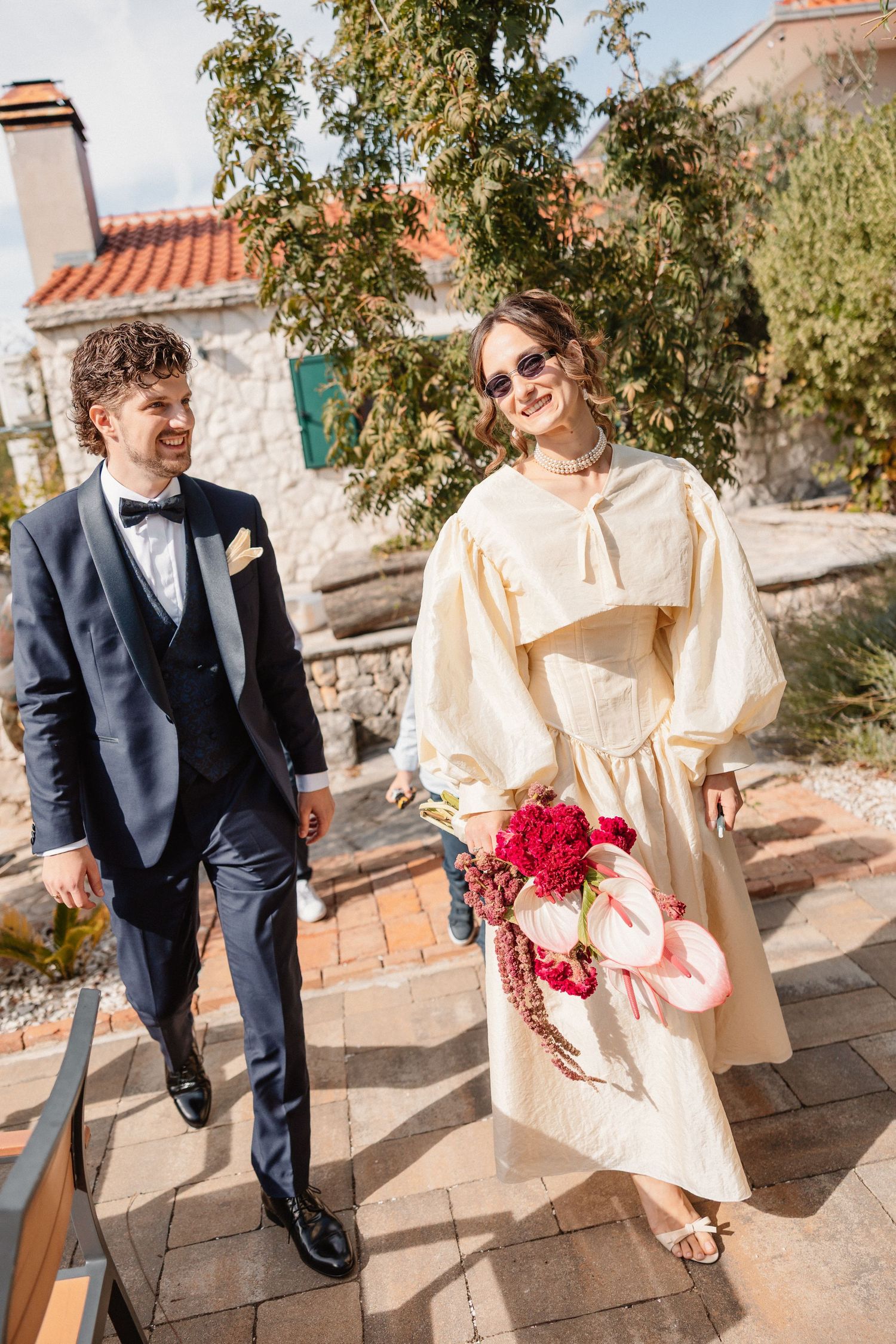 Couple walking on stone path, one in navy suit and other in cream dress with red flowers.