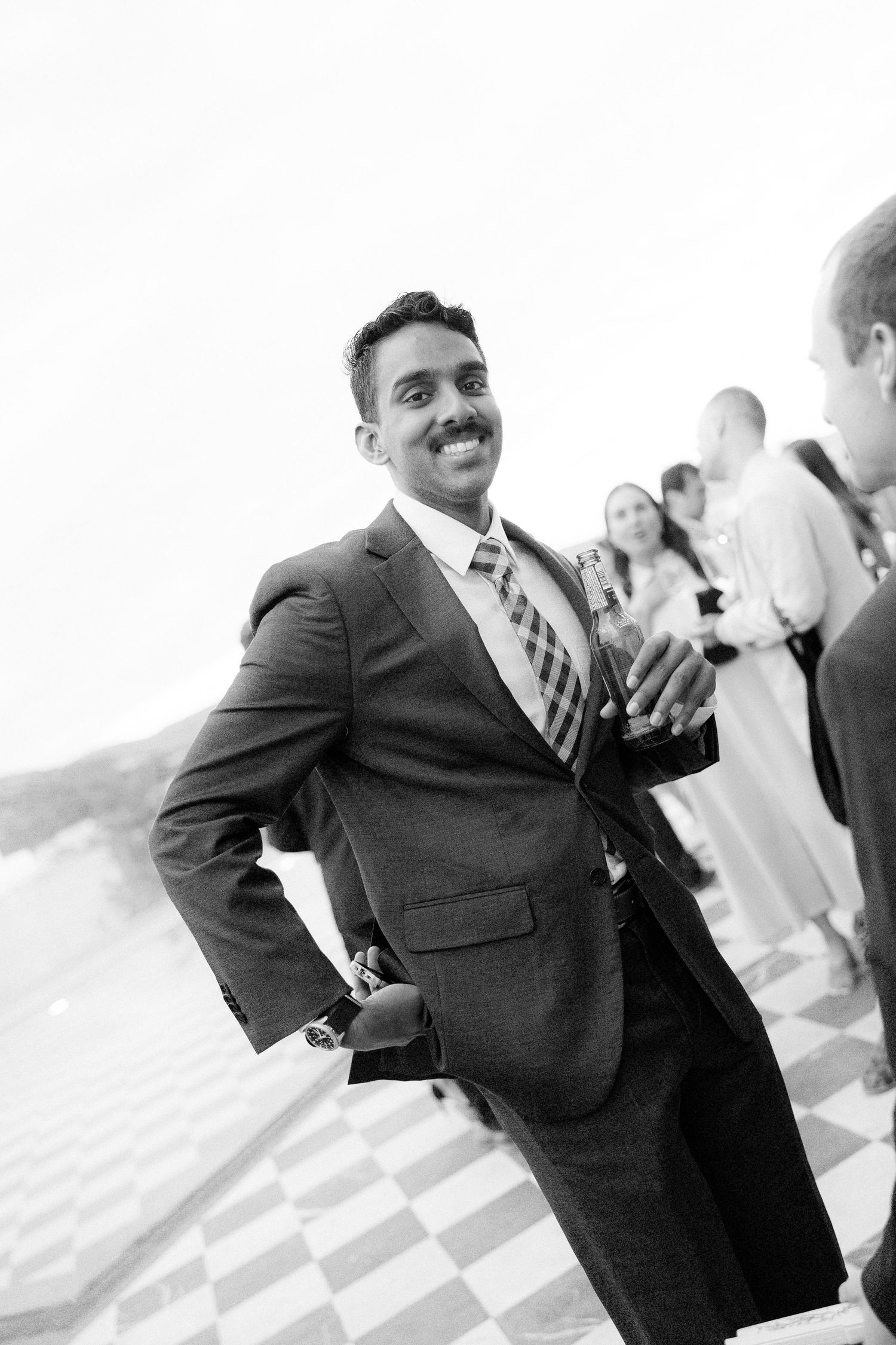 Smiling man in gray suit and striped tie poses at waterfront wedding reception in black and white.