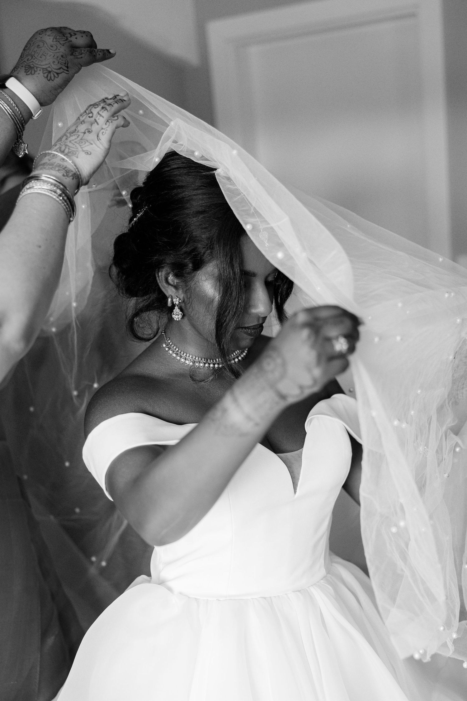 Black and white photo of bride in strapless gown having veil placed over her head by henna-adorned hands.
